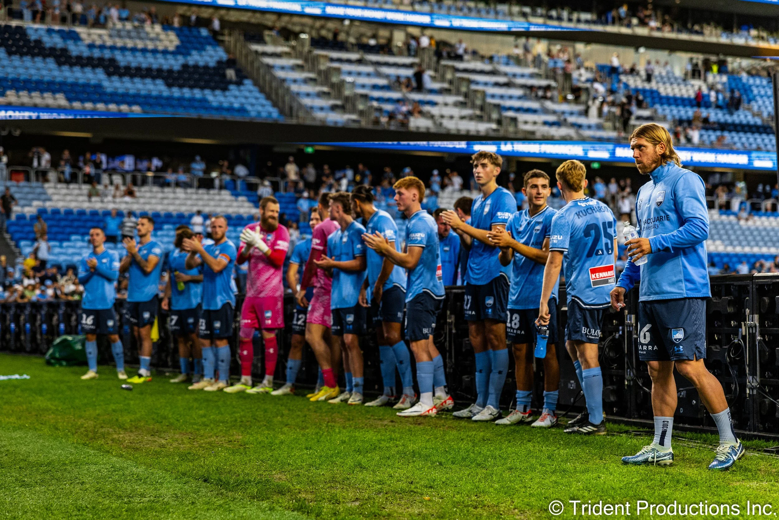 Soccer team standing in a line on the sideline of a stadium, clapping before a game, wearing blue jerseys and shorts, with some players holding water bottles.