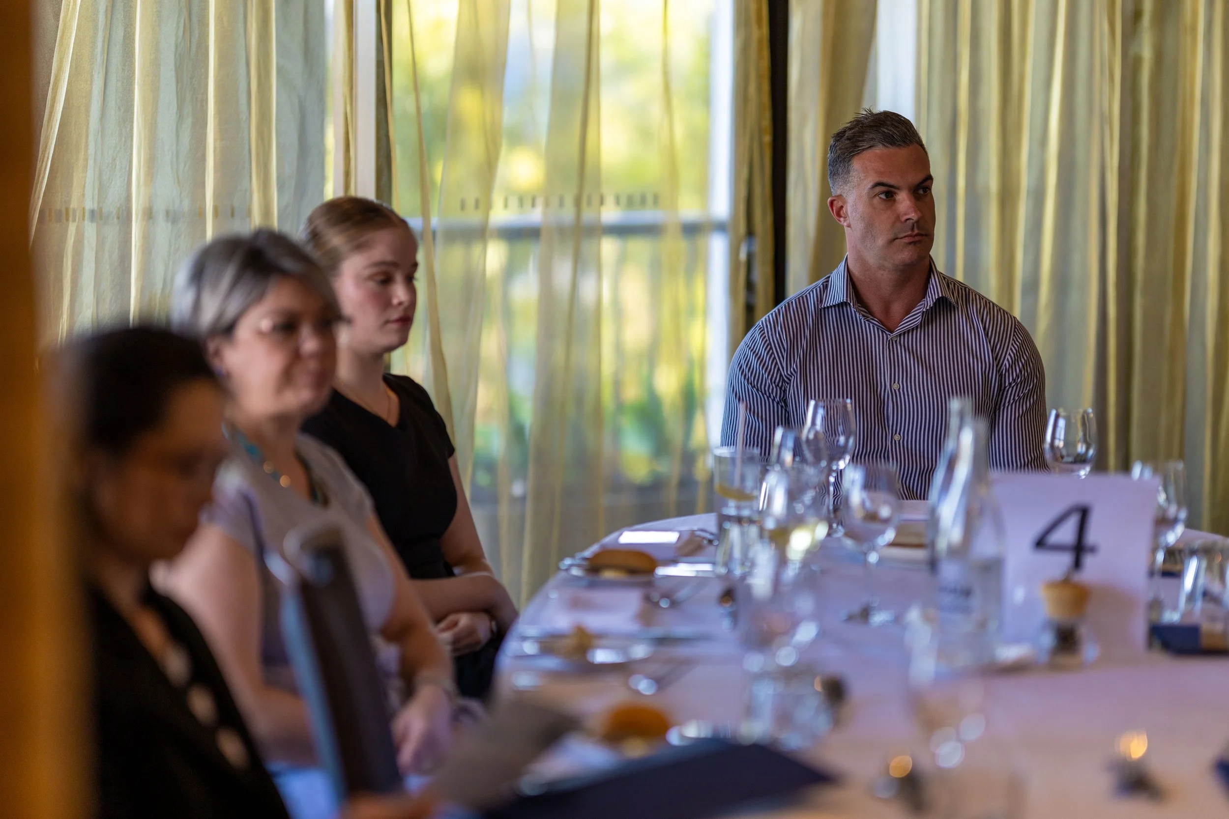 People sitting at a banquet table during a meeting or event, with a man in a striped shirt in focus on the right and three women blurred in the foreground on the left, in a room with yellow curtains and large windows.