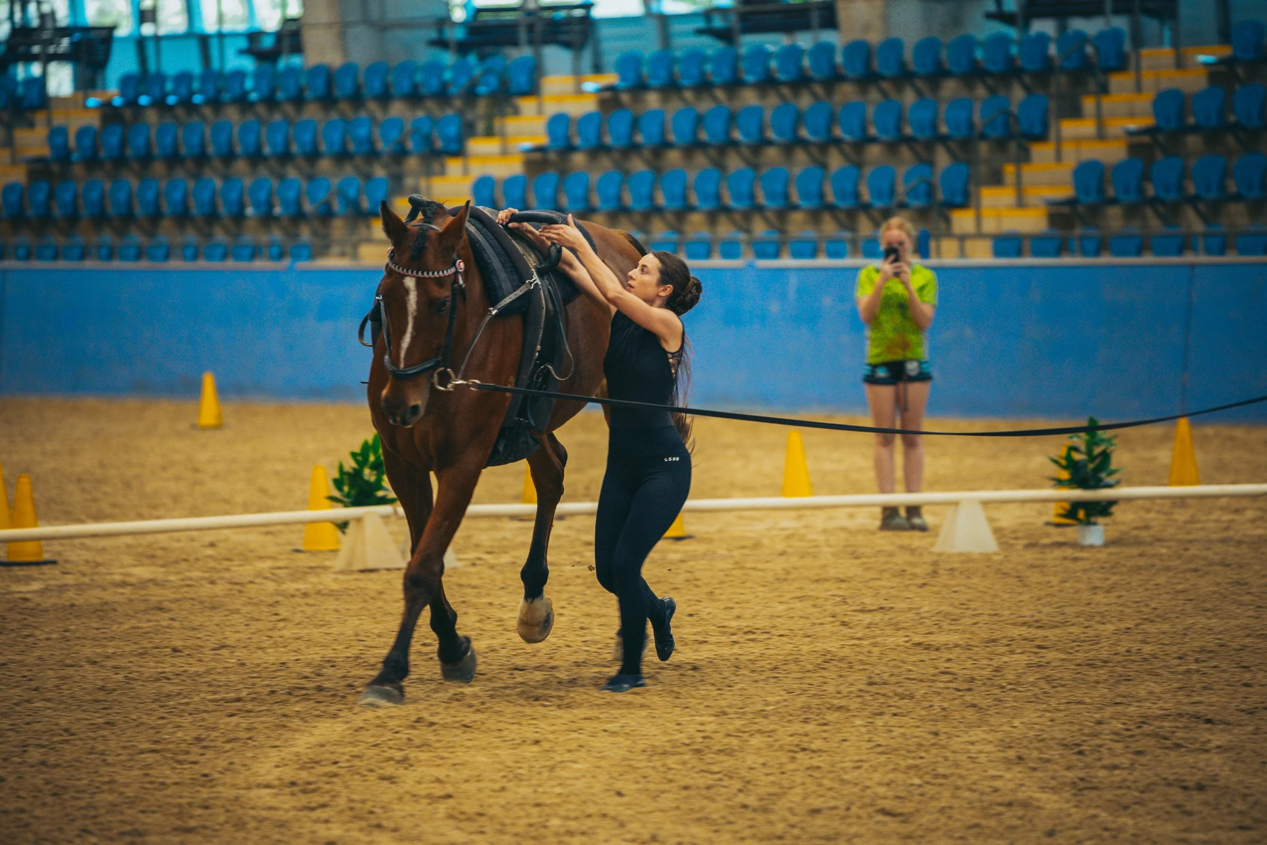 A woman in athletic clothing adjusts a saddle on a brown horse in an indoor riding arena, with another woman in a neon green shirt taking photos in the background.