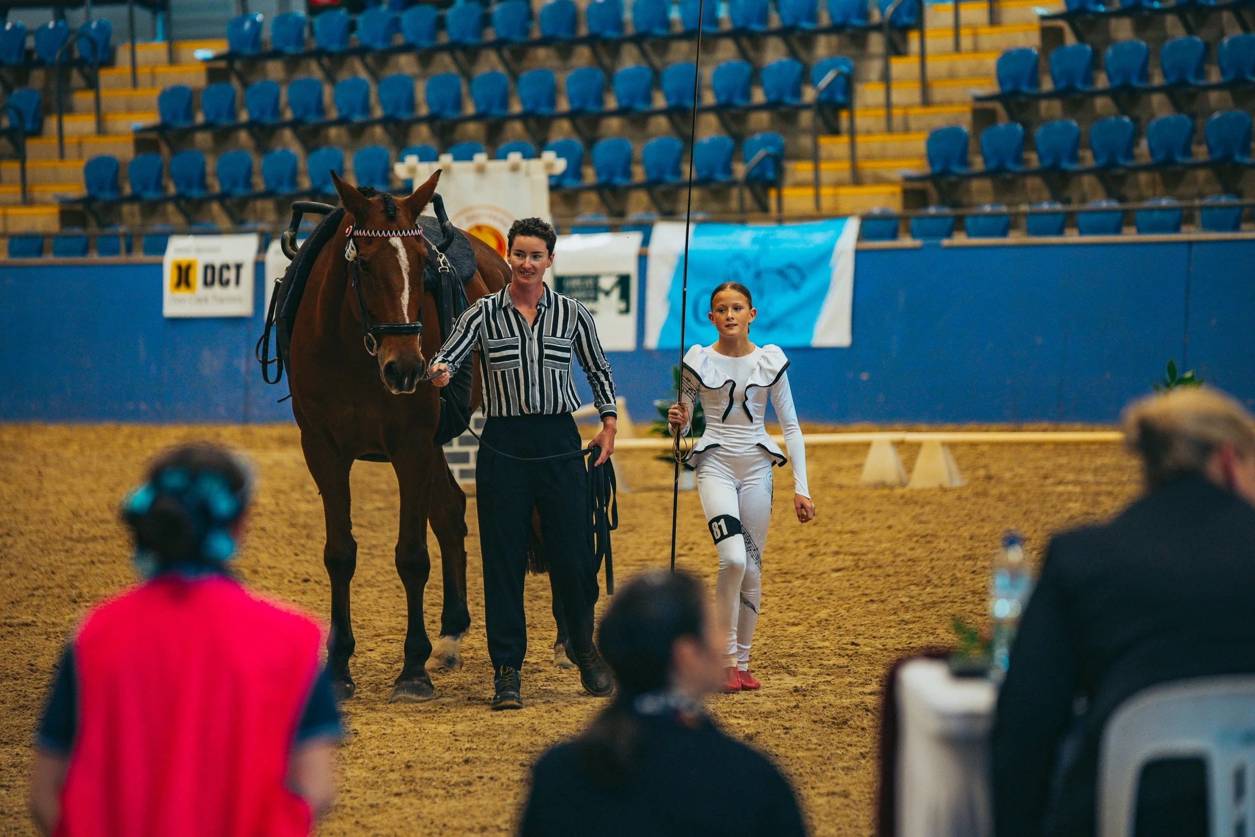 A woman leading a brown horse in an indoor arena while a young woman in a white costumes with the number 81 walks beside her holding a whip. Spectators or judges are seated in the foreground.