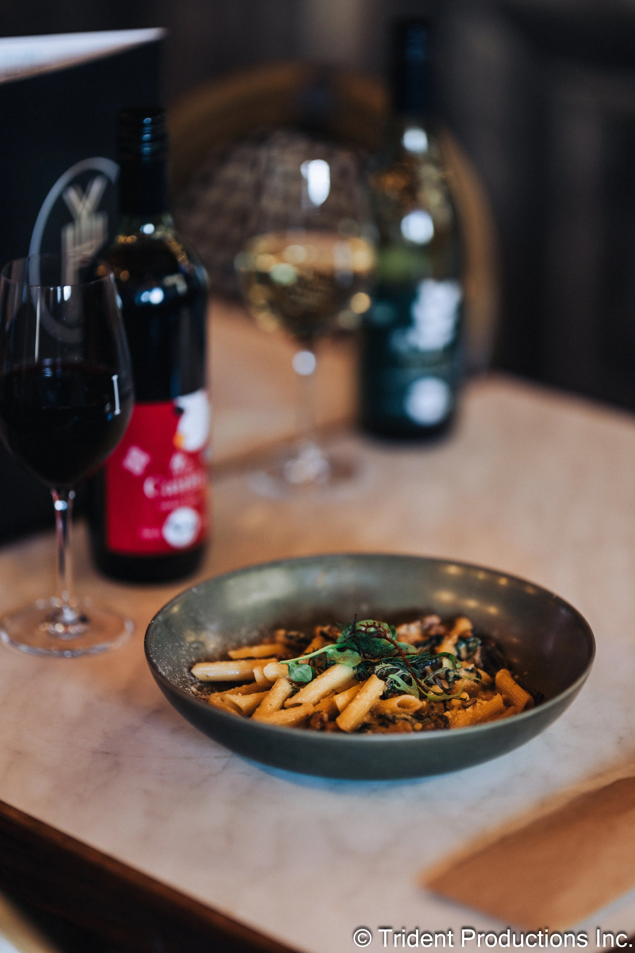 A bowl of pasta garnished with microgreens. In the background, there is a glass of red wine, a bottle of wine, and some blurred decorative items.