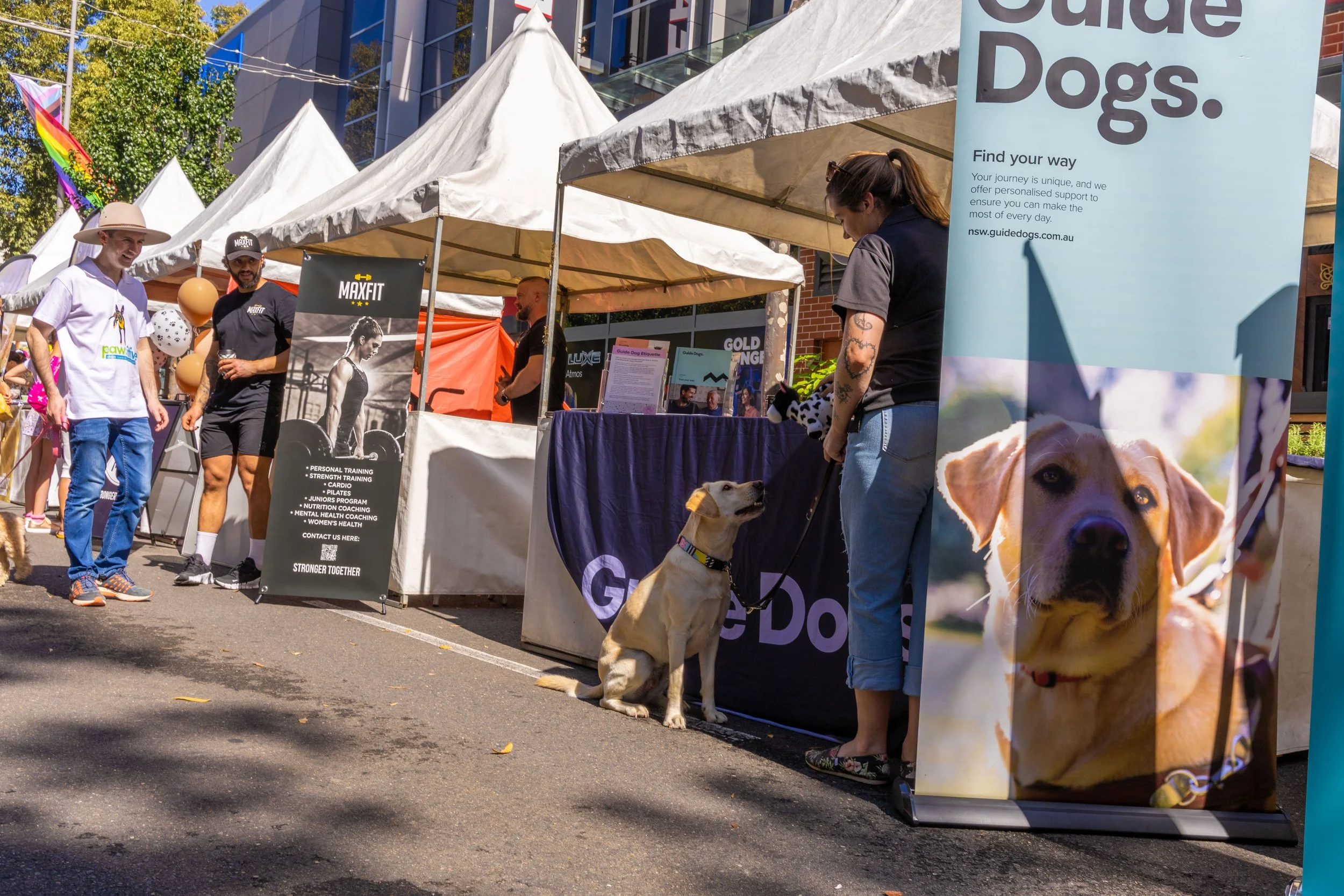 Outdoor event with tents and booths, including a dog guide service booth with a woman and her service dog, people walking and talking, some holding dogs, and various promotional banners.