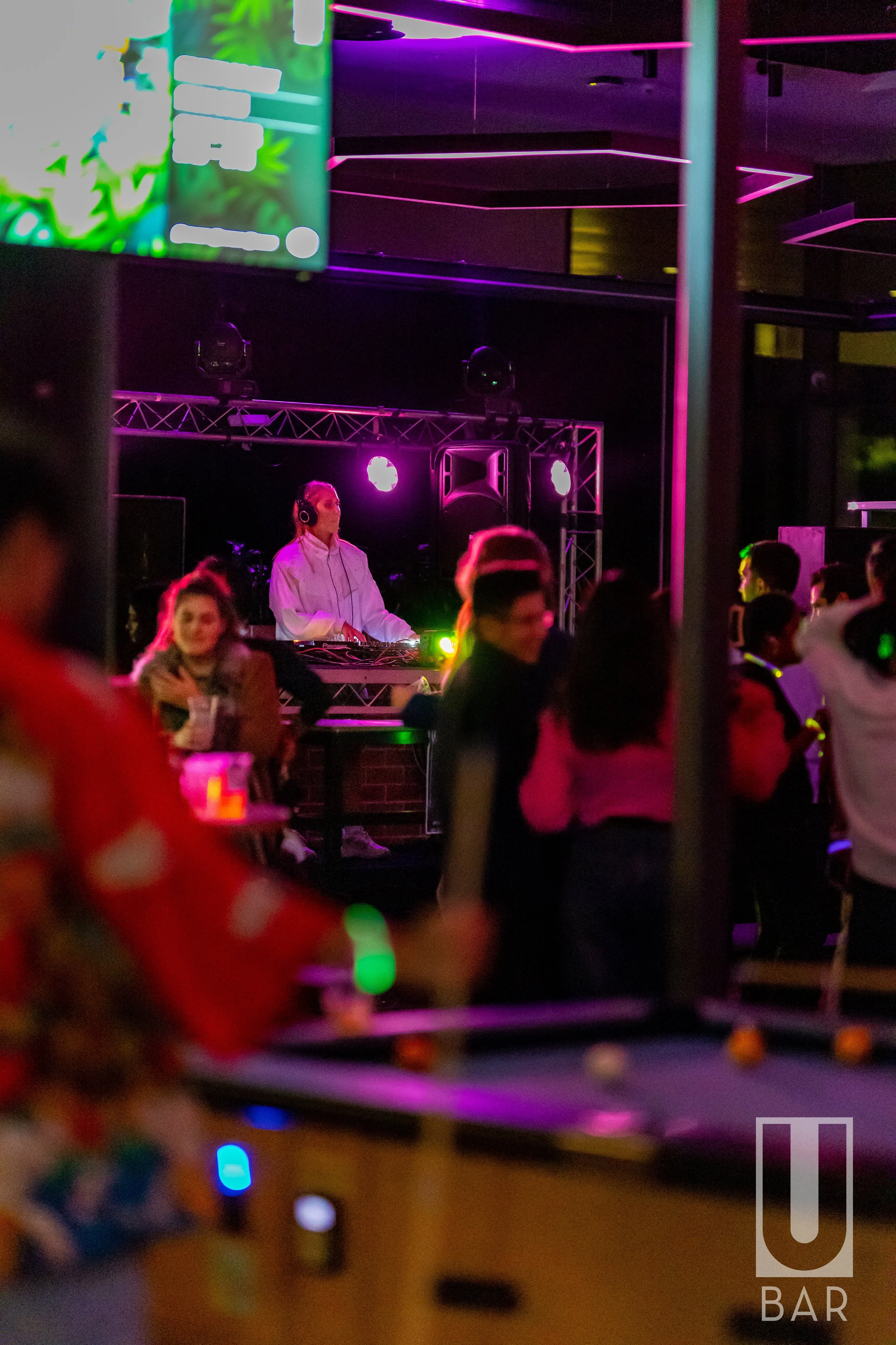 A DJ wearing headphones and a white shirt performing on stage at a nightclub with pink purple lighting. People dancing around, and a foosball table in the foreground.