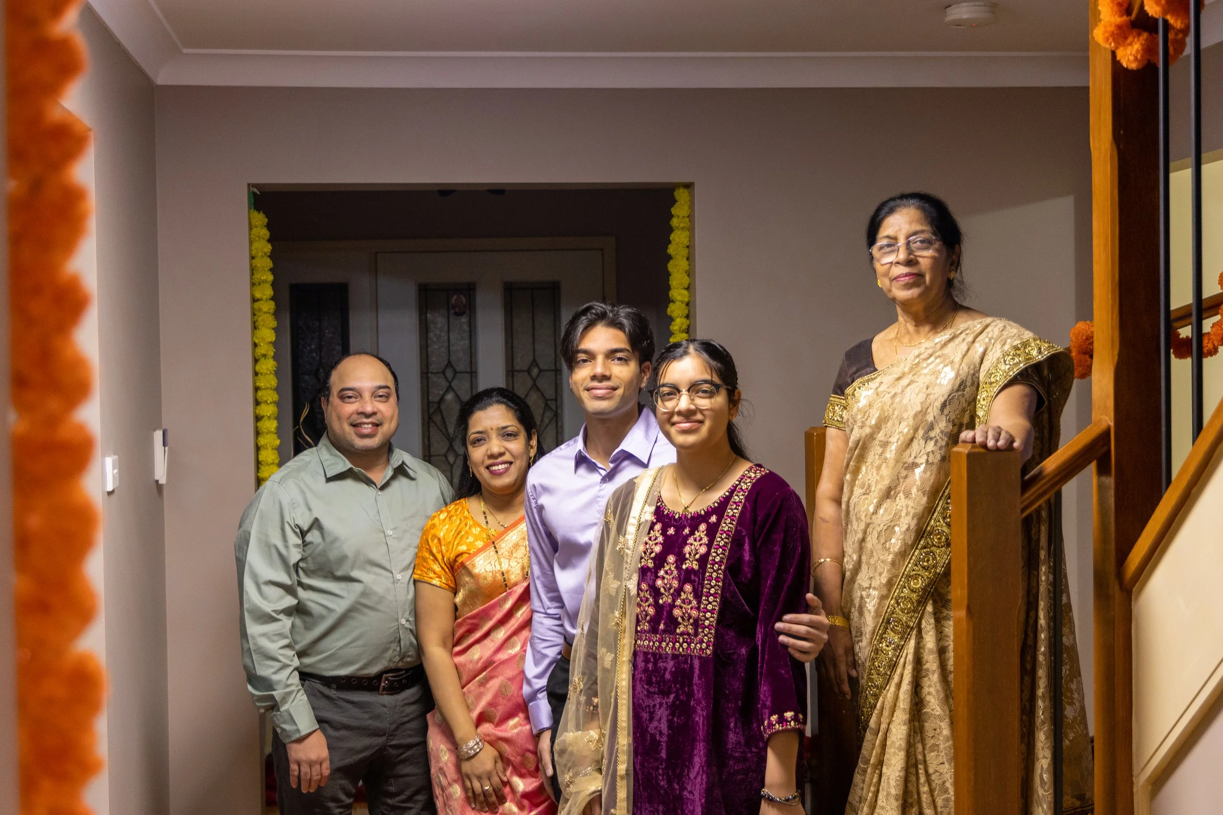 A multi-generational Indian family celebrating indoors. They are dressed in traditional Indian attire, standing on and near a staircase decorated with marigold flowers.