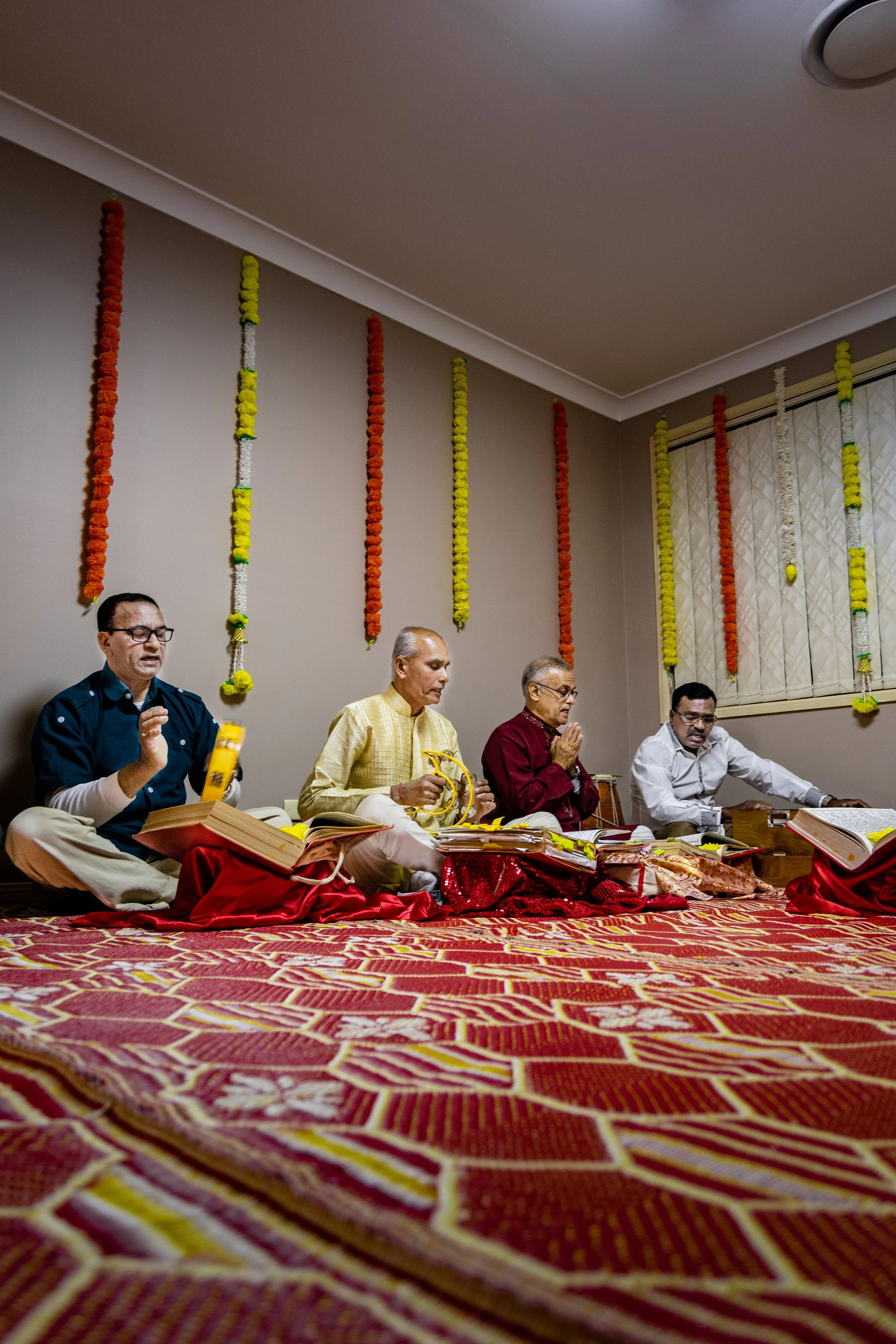 Five men sitting on the floor during a religious ceremony or prayer session, with books and flowers nearby, decorated with flower garlands on the wall.