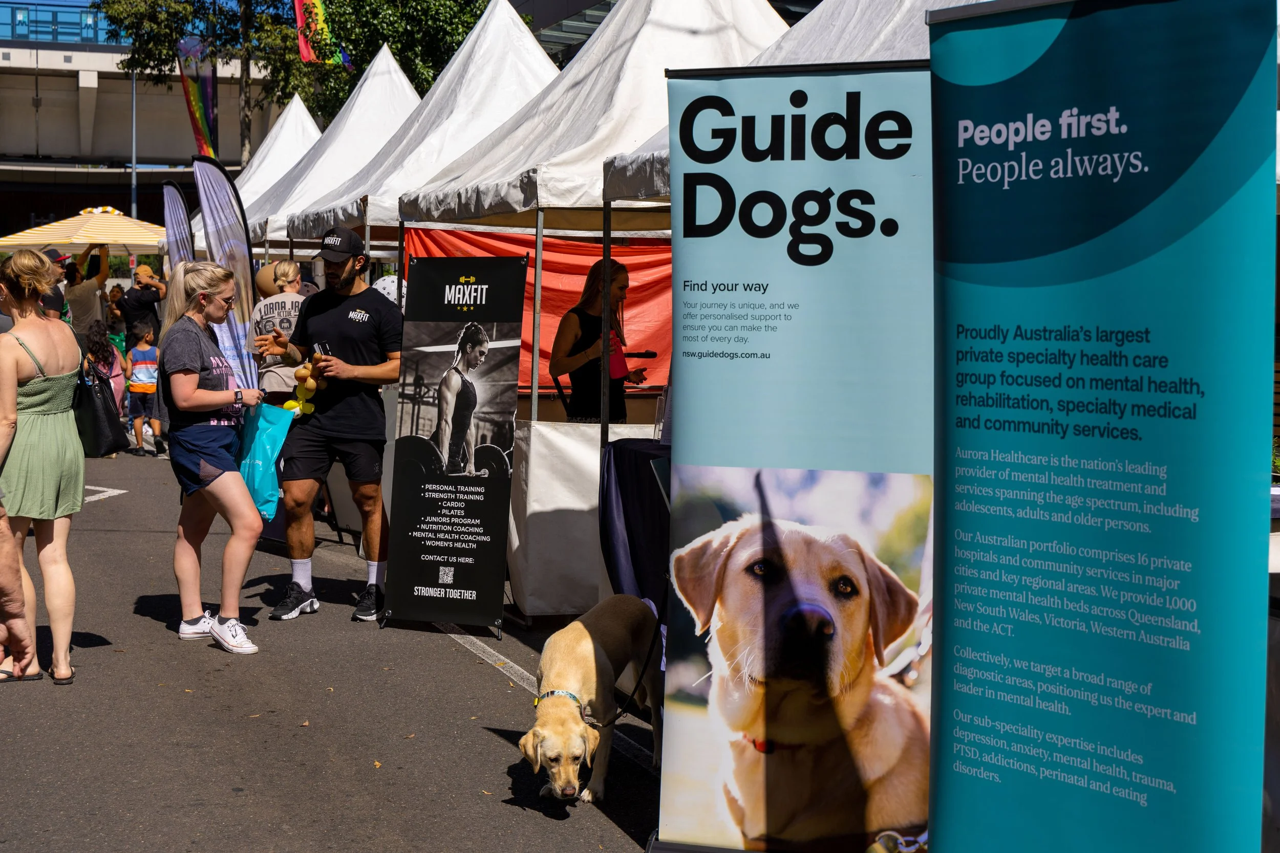 People attending an outdoor event with white tents, including a booth for Guide Dogs with a large poster of a dog, and a man in black attire talking to a woman. Other signs and booths are visible, and there is a yellow dog on a leash near the Guide D
