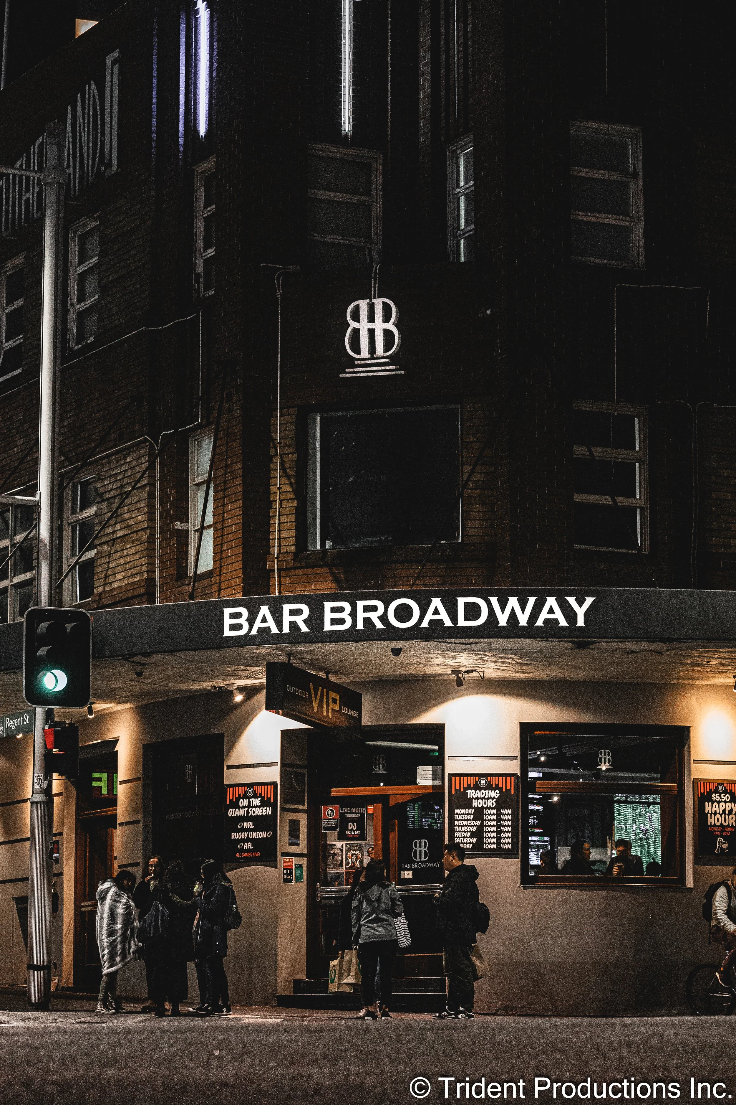 Nighttime exterior view of Bar Broadway with people gathered outside, illuminated signs, and a traffic light in the foreground.
