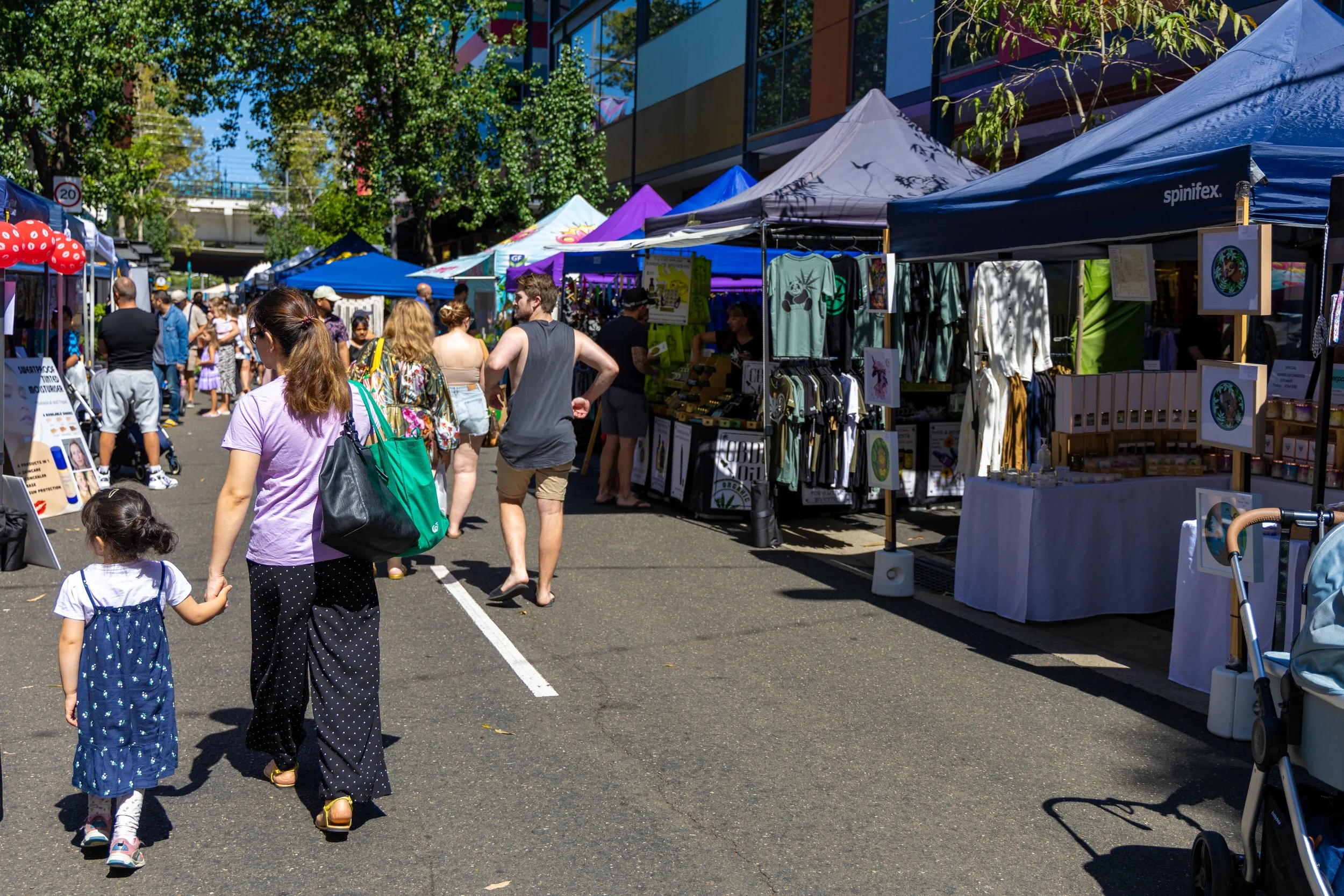 Outdoor street market with vendor tents and people shopping on a sunny day, with trees and buildings in the background.