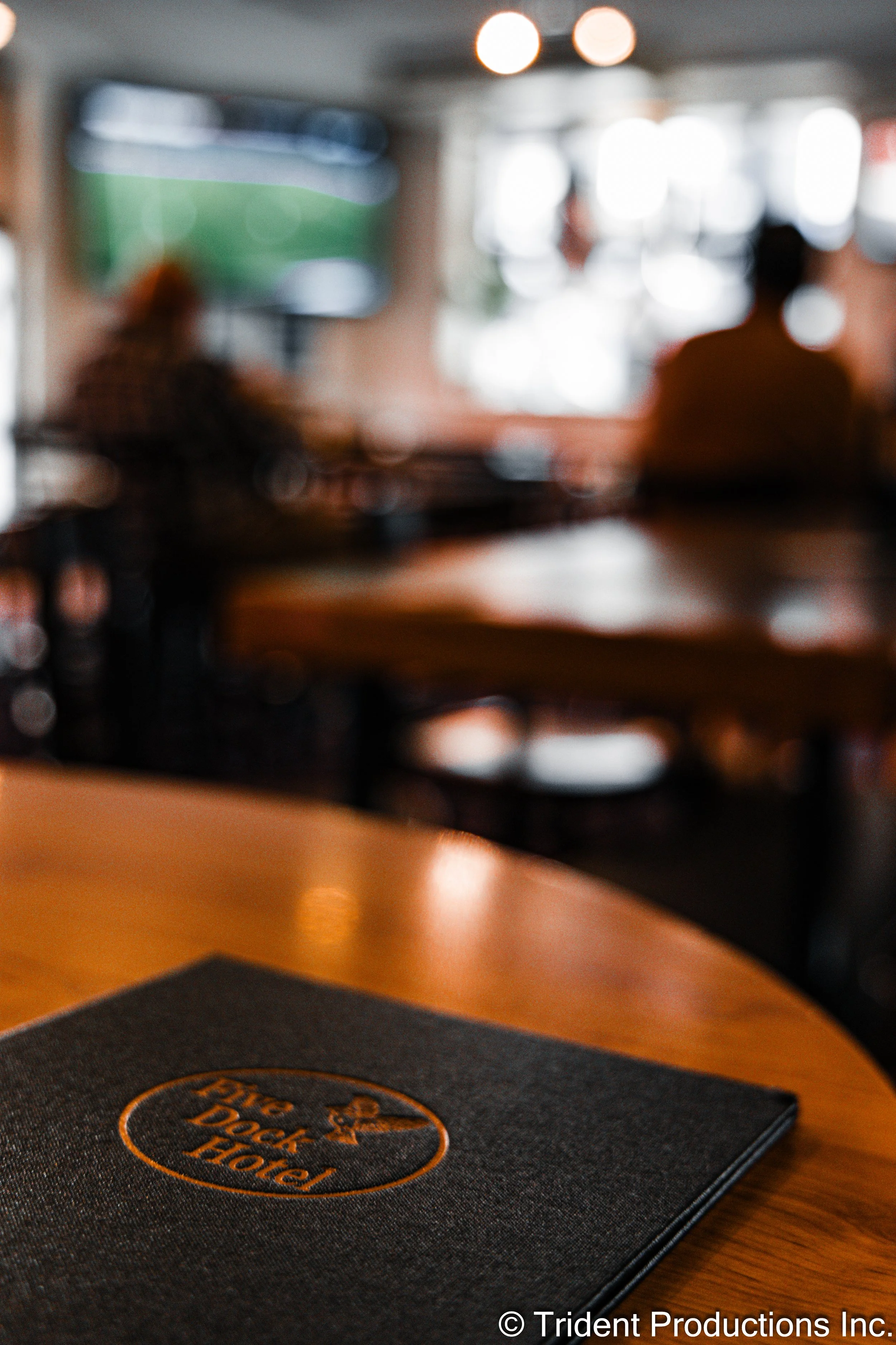 A menu or napkin from Big Rock Hotel on a wooden table inside a restaurant, with a television and blurry figures of people in the background.