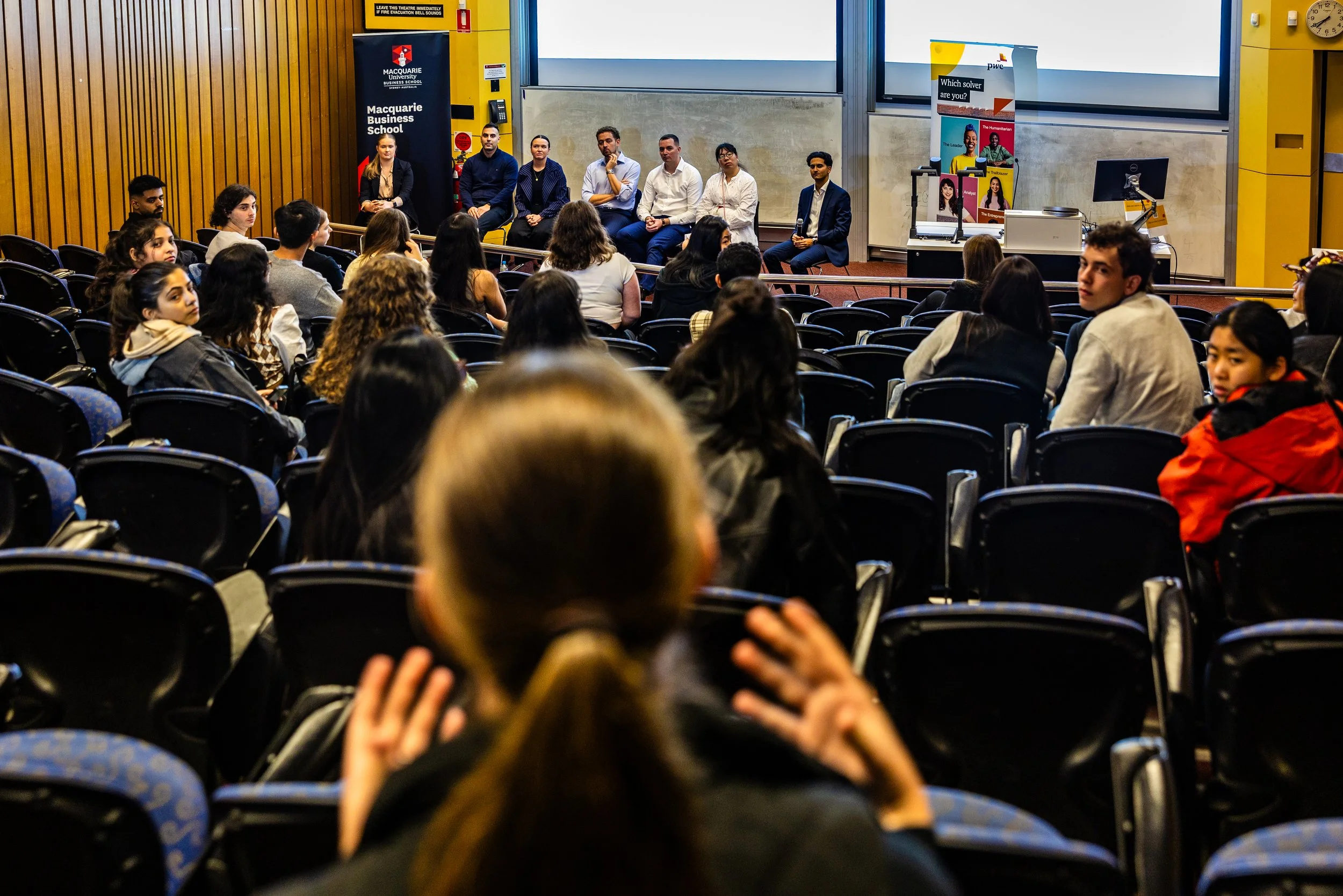 A lecture hall with students sitting in chairs, facing a panel of speakers at the front of the room.