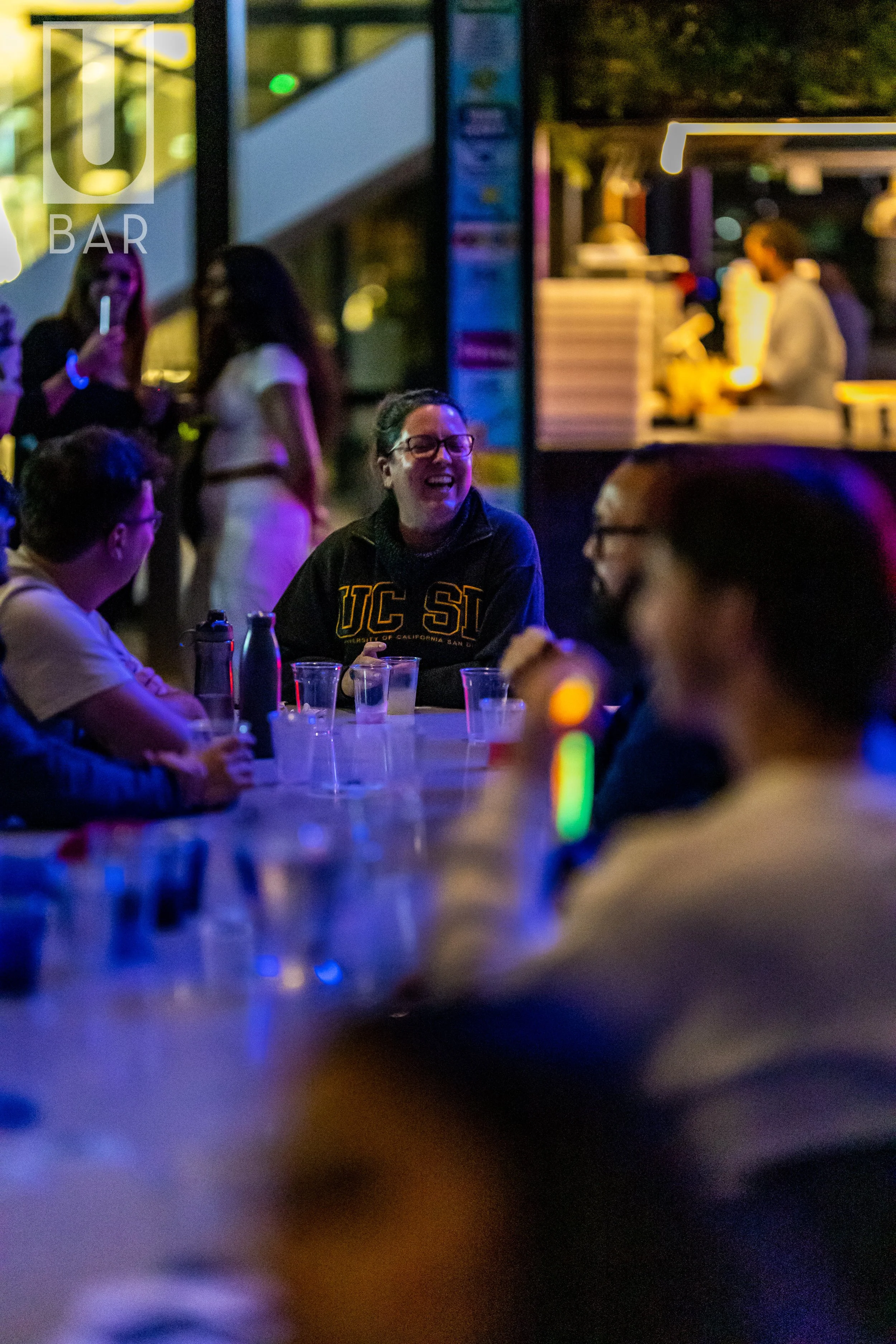 People gathering at a bar or restaurant during the evening, with some individuals sitting at a table and others standing. The scene is illuminated with colorful, dim lighting, creating a lively atmosphere.