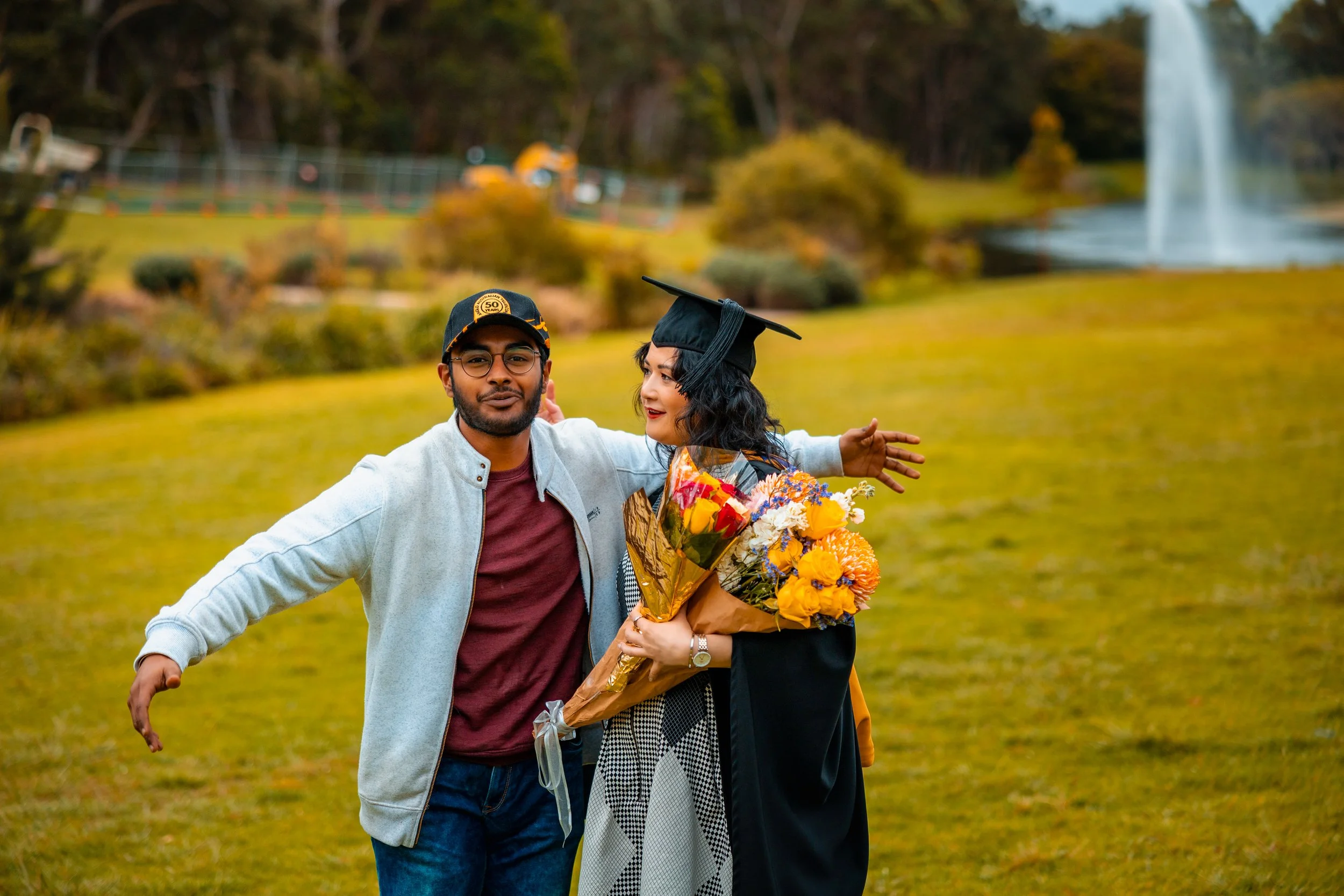 A young woman in a graduation cap and gown holding a bouquet of flowers celebrates her graduation outdoors, with a man smiling beside her on a grassy field and a fountain in the background.