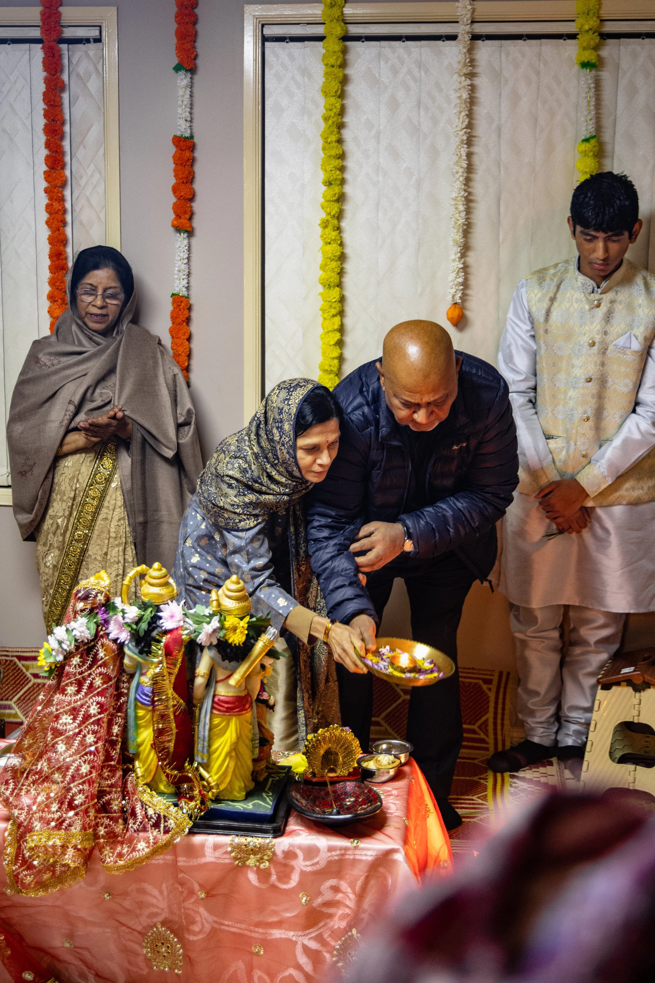 People participating in a Hindu religious ceremony, with statues of deities and offerings on a decorated table.