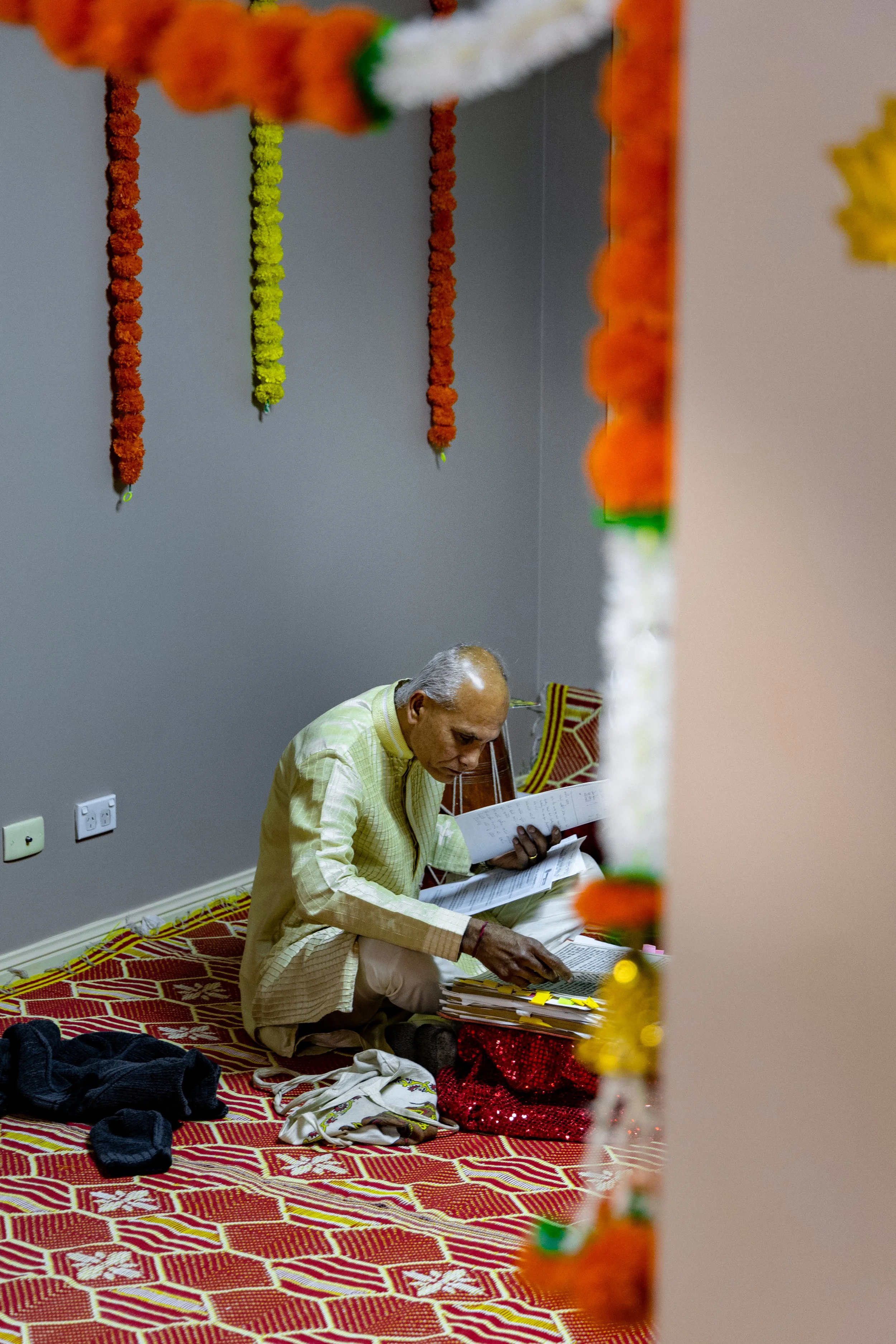 An elderly man sitting on a colorful patterned carpet, reading a book with papers and a red sequined bag nearby, seen through an ornate doorway decorated with marigold flower garlands.