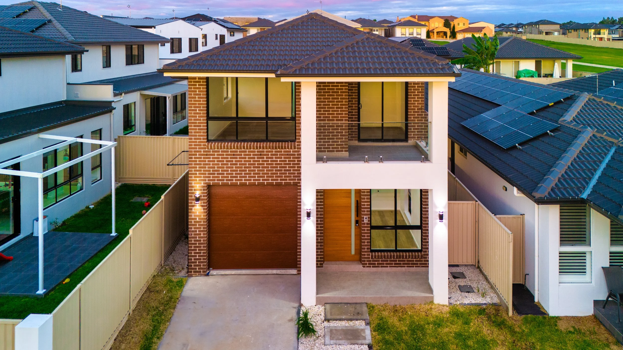 A modern two-story house with a brick and white exterior, large glass windows, and a balcony on the upper floor. The house features a brown tiled roof and is situated in a suburban neighborhood with other similar houses. There is a paved driveway lea