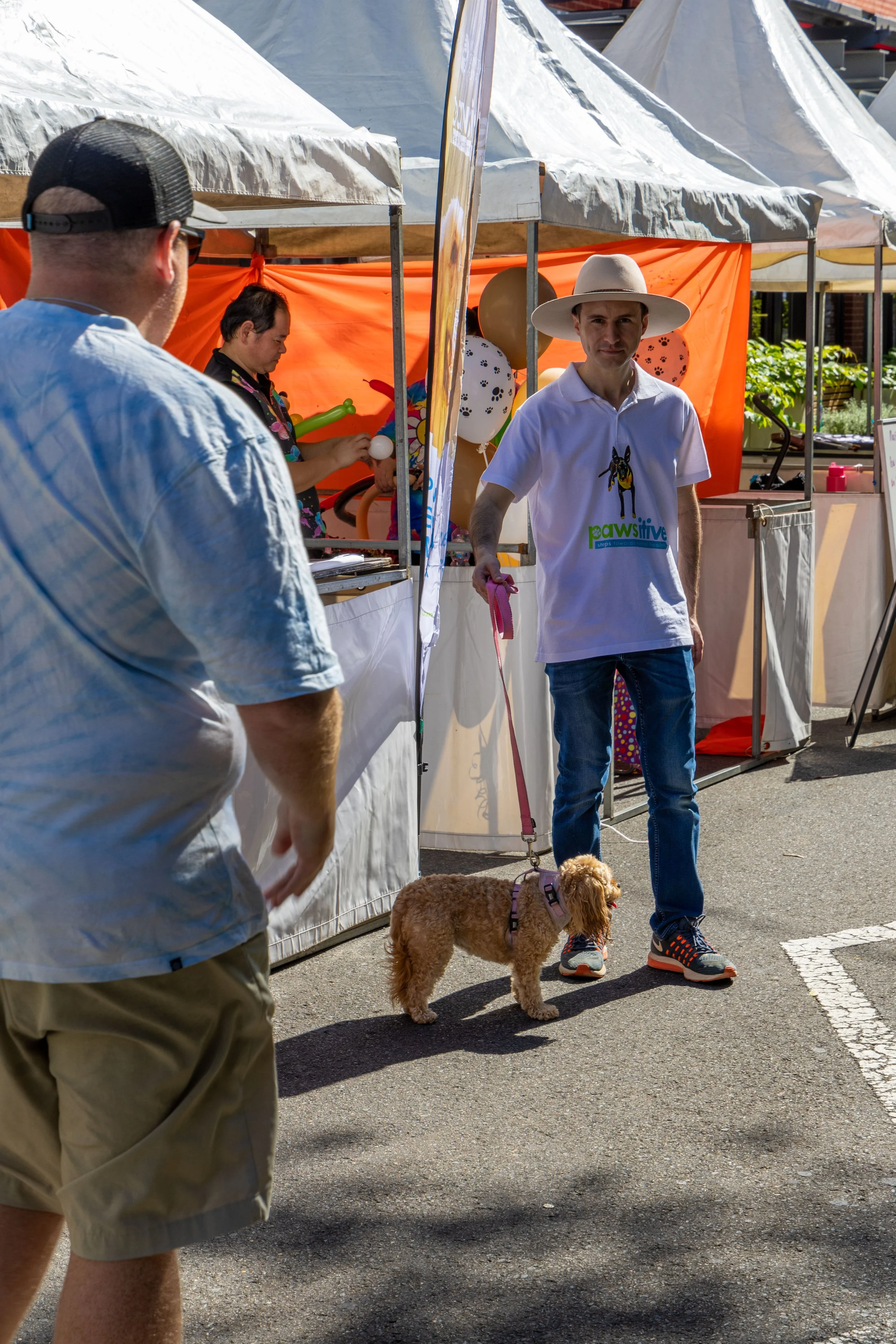 A man holding a dog on a leash in front of a booth at an outdoor fair or market, with another person in the foreground. The booth has a bright orange backdrop and balloons, and is covered with beige and white tents. The man is wearing a white hat, a 