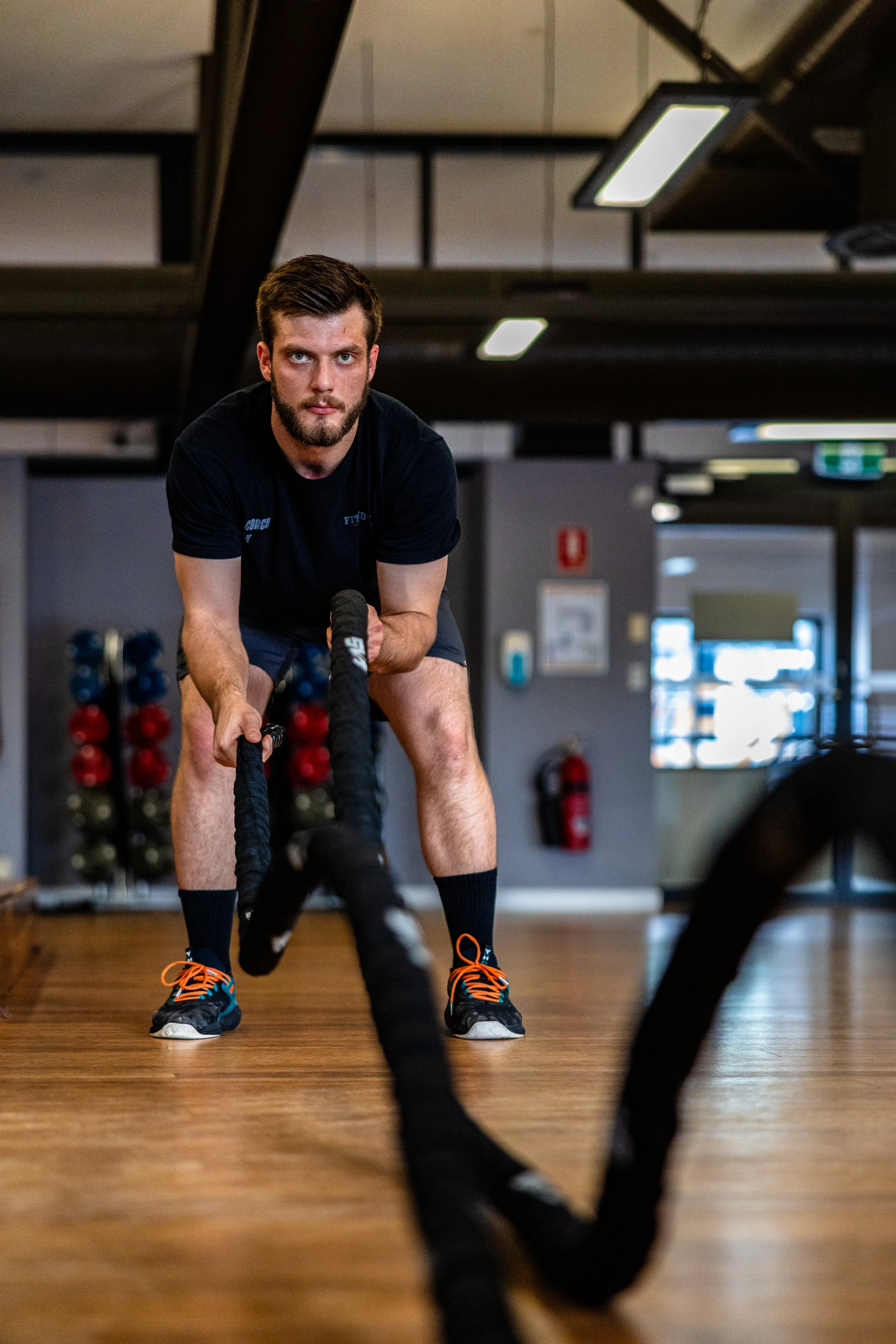 Man in black shirt and shorts exercising with battle ropes inside a gym.