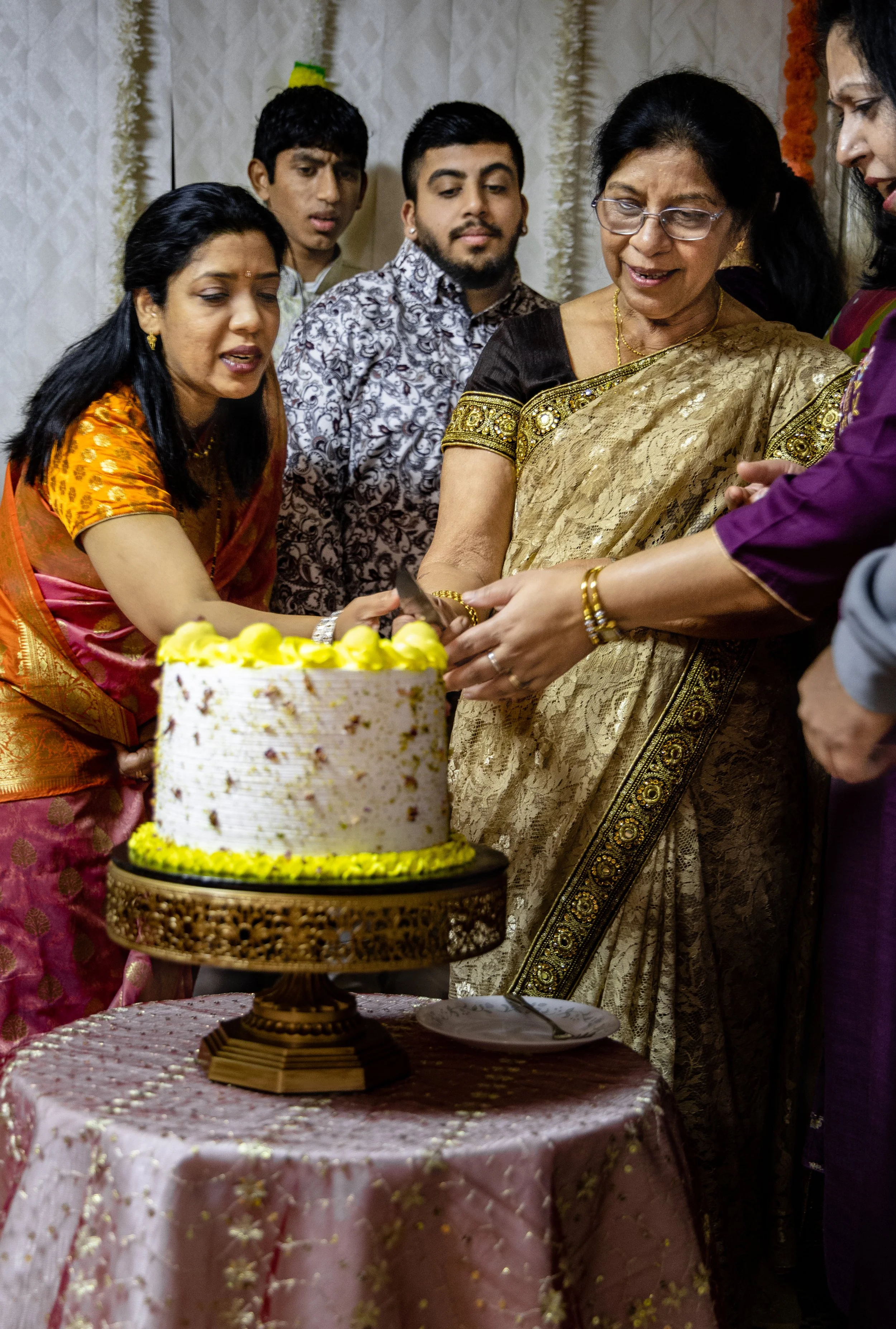 Group of people celebrating around a birthday cake, with a woman in traditional Indian attire cutting the cake.