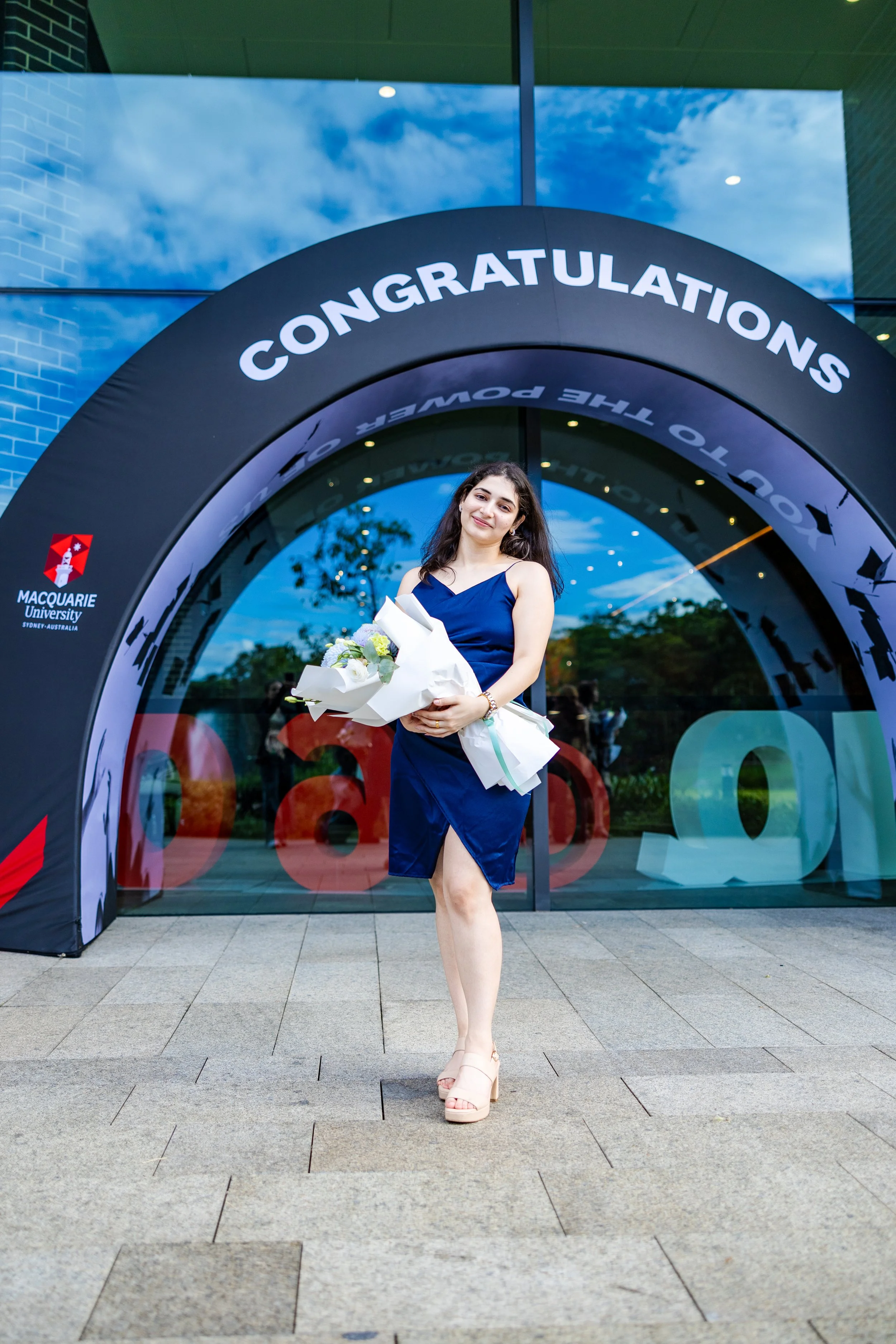 A young woman in a blue dress holding a bouquet of flowers standing in front of an archway that says 'Congratulations' at Macquarie University in Sydney, Australia.