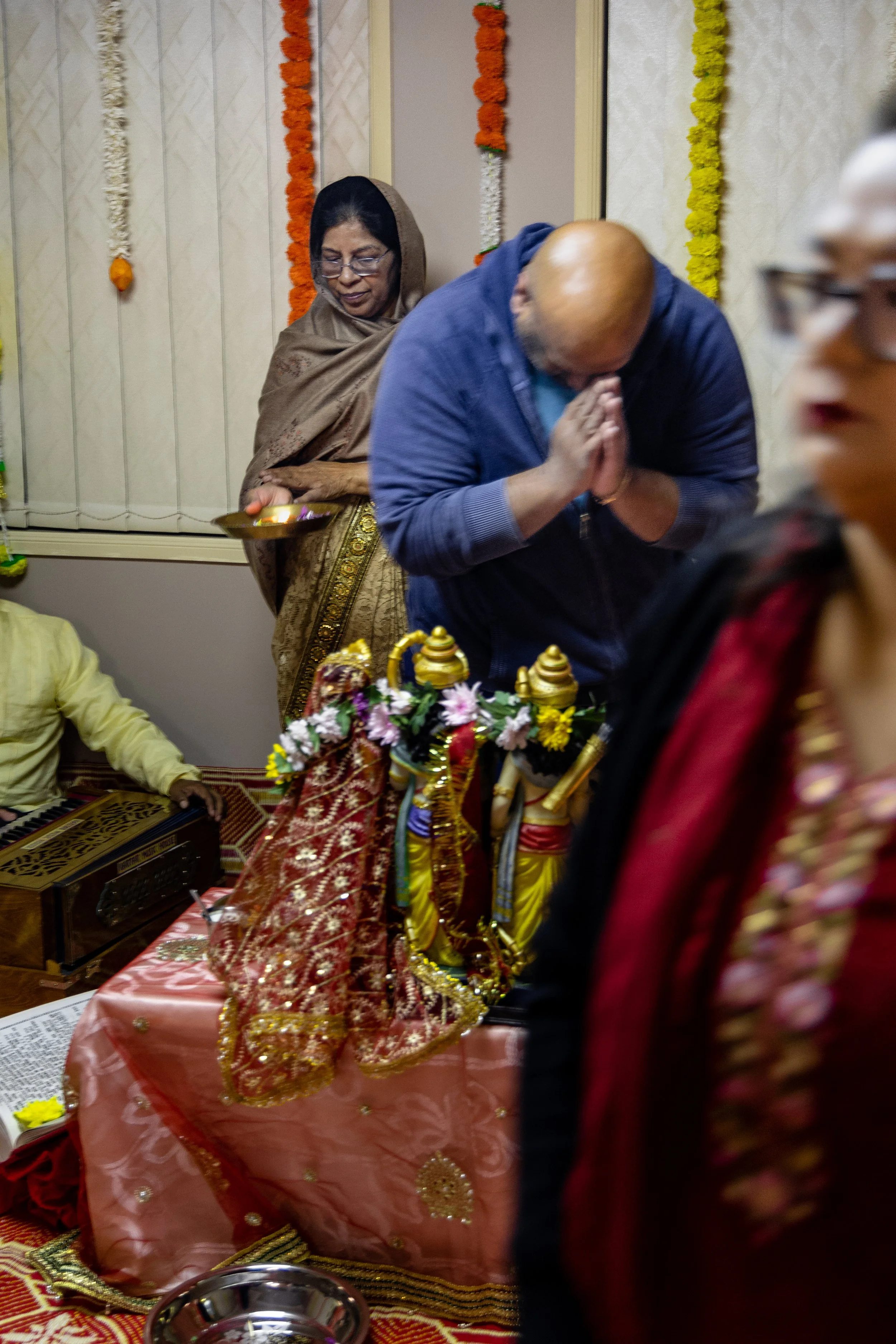 People participating in a Hindu religious or cultural ceremony, with a woman in traditional attire and devotion, and a man with folded hands in prayer, in front of decorated idols and flowers.