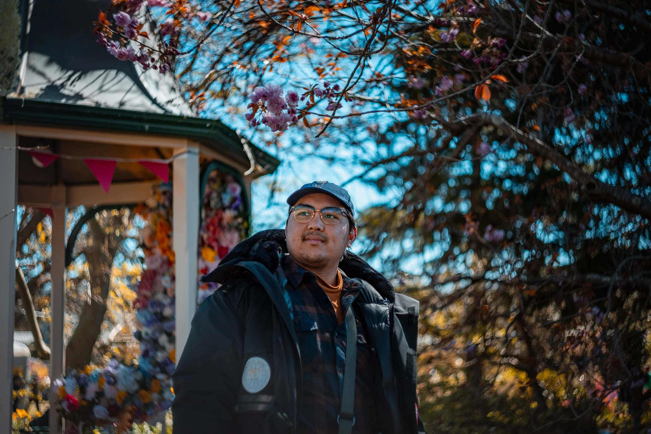 A man wearing glasses, a dark jacket, and a hat standing outdoors with autumn-colored trees and a decorated structure in the background.