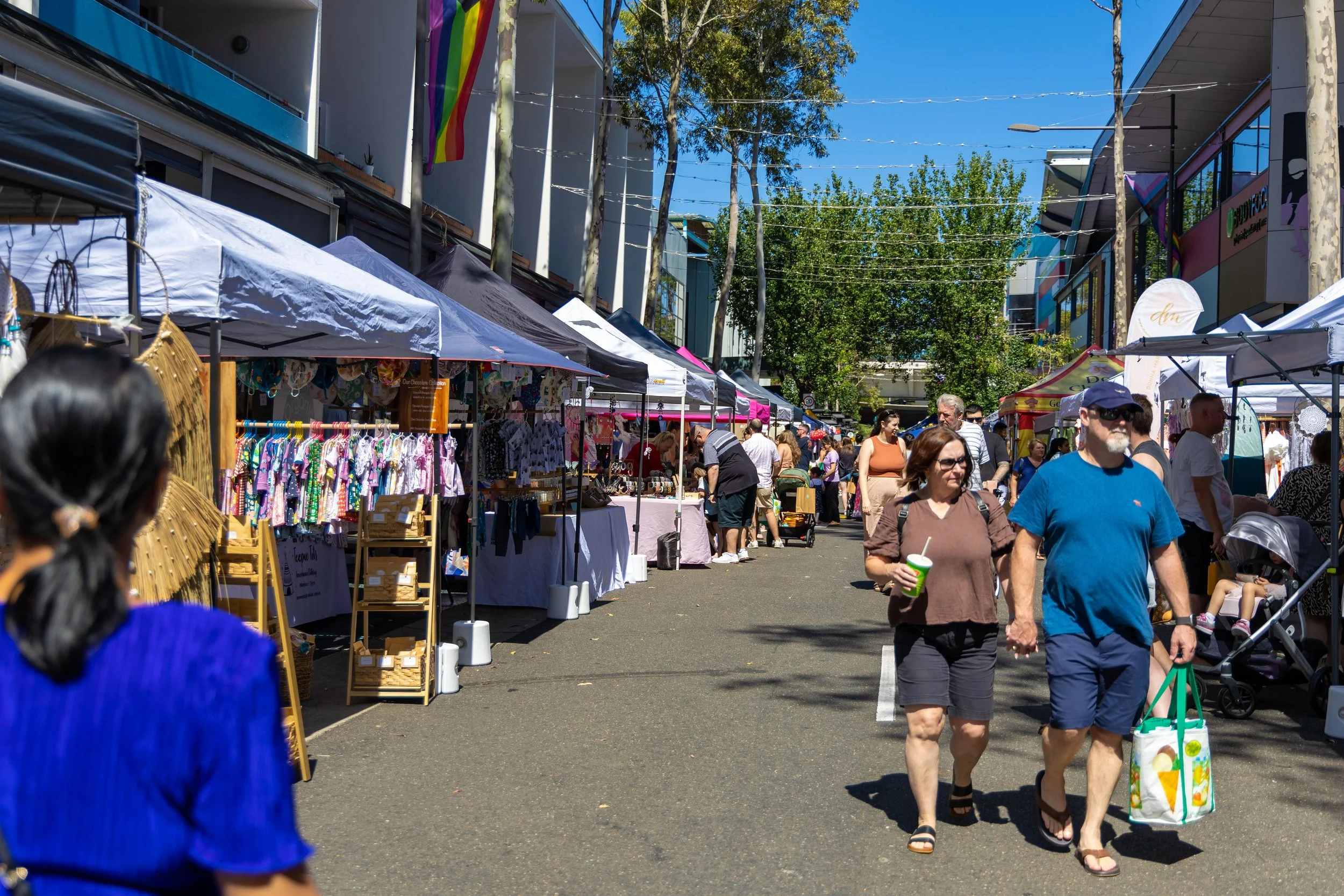 People walking and browsing at an outdoor street market with vendor tents on both sides. The market is set up on a sunny day in an urban area with trees and modern buildings in the background.