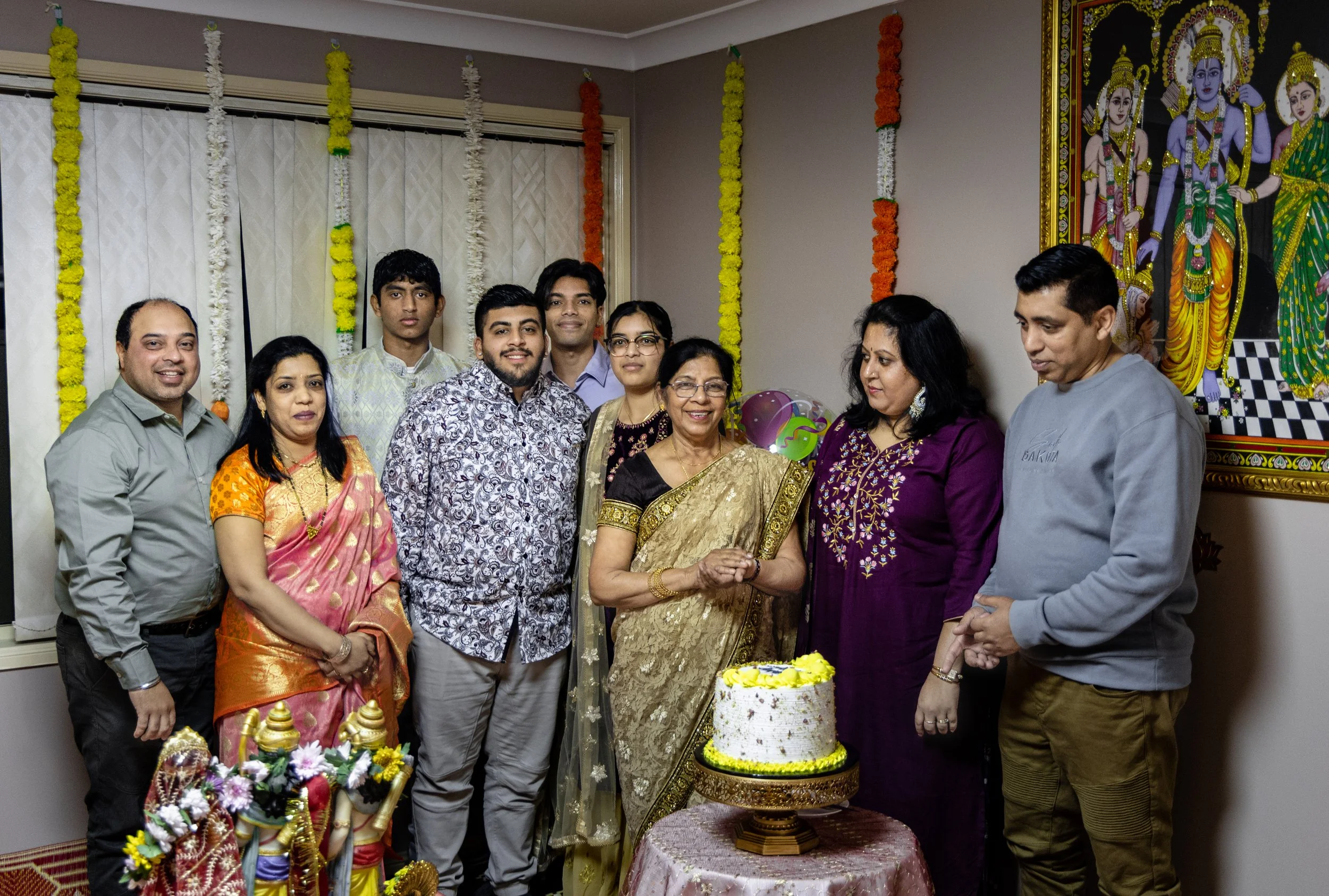 A group of people celebrating an Indian religious or cultural event indoors. They are standing around a decorated cake and religious icons, with traditional attire, floral decorations, and a large colorful painting of Hindu deities on the wall.