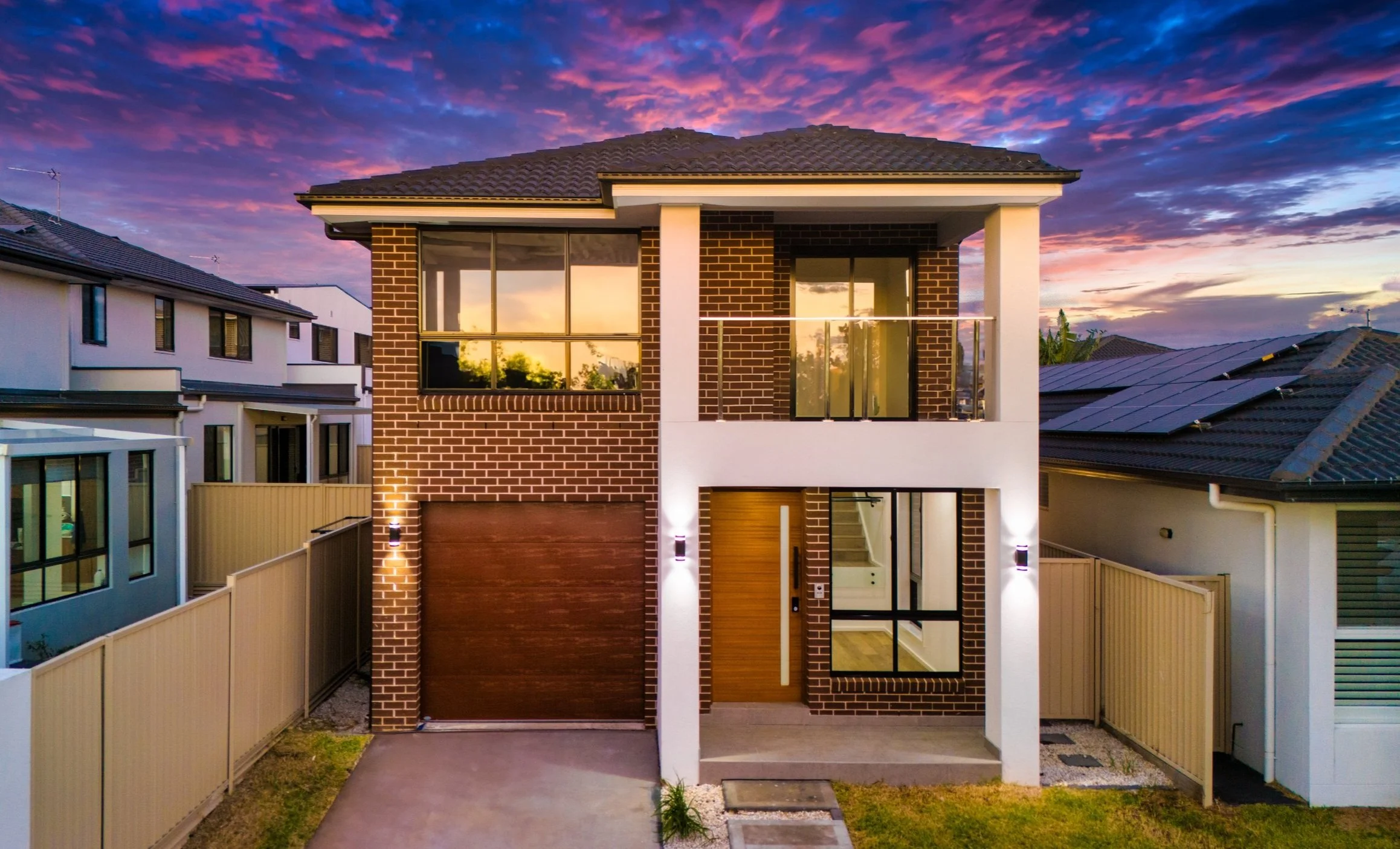Modern two-story house with a brick facade, large windows, and a wooden front door, illuminated at sunset with a colorful sky in the background.