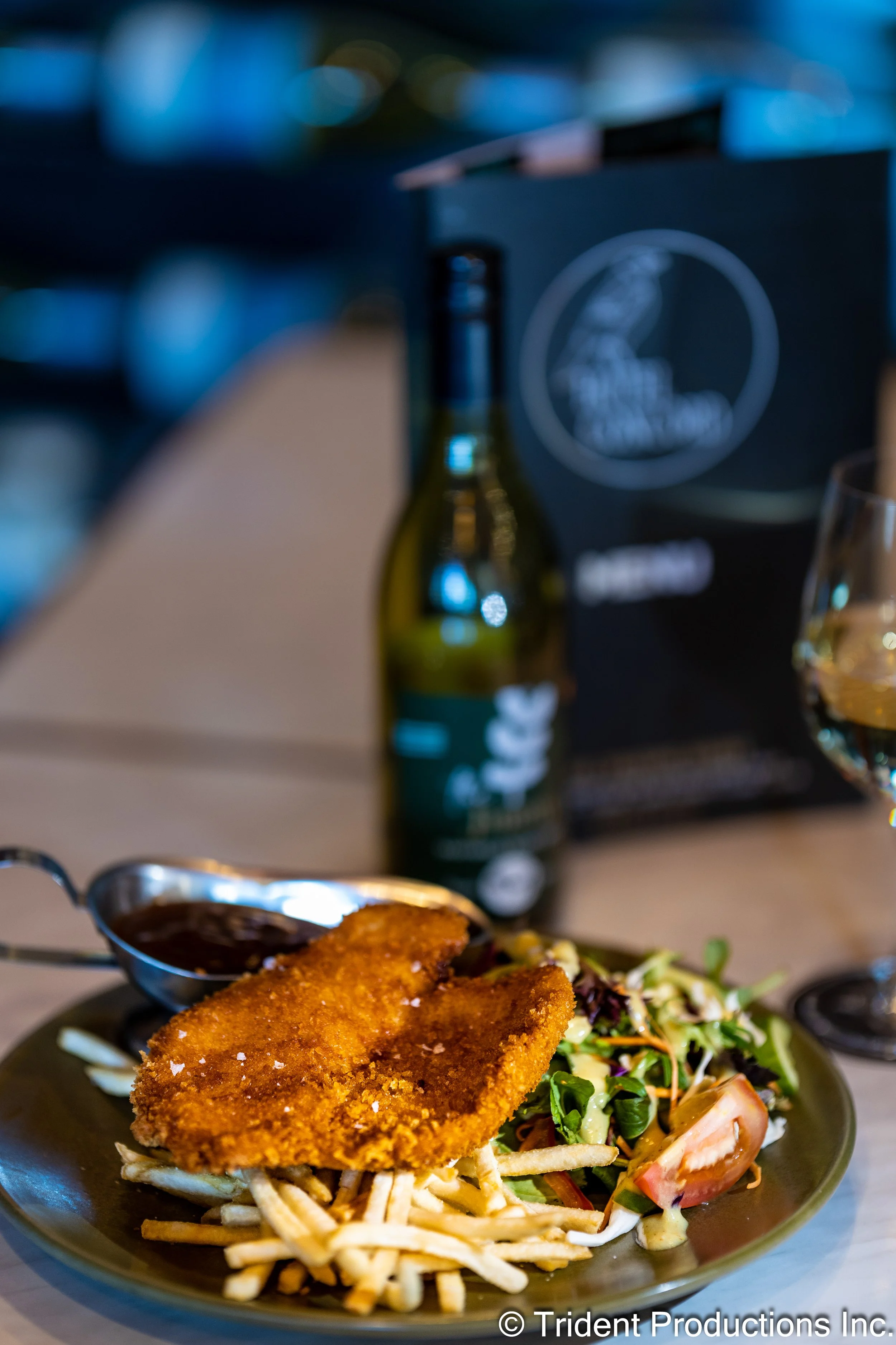 Plate with breaded fish fillet, French fries, and salad with lettuce, tomato, and dressing, with a spoon of dark sauce, in a dining setting.