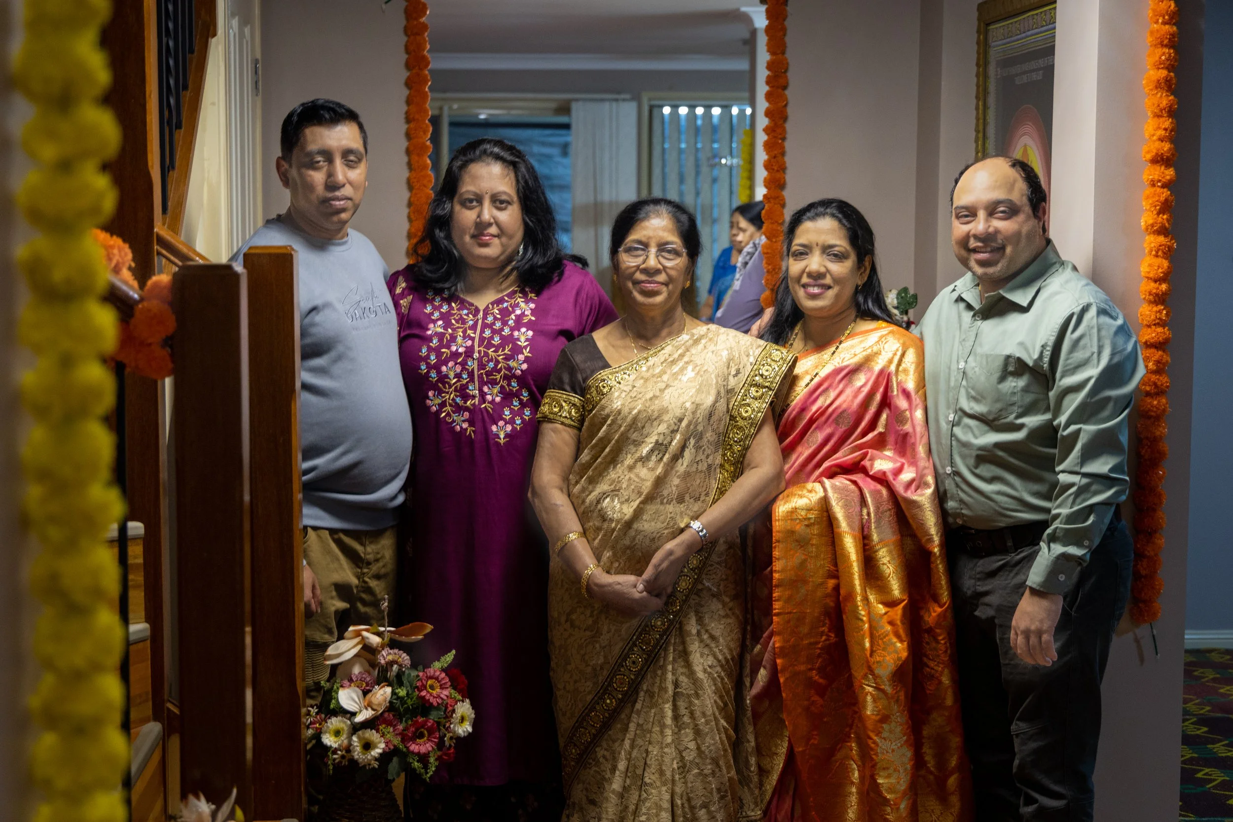 Group of five people standing together inside a decorated room, posing for a photo. Three women are wearing traditional Indian dresses, and two men are wearing casual shirts. The background shows a doorway with a floral garland.