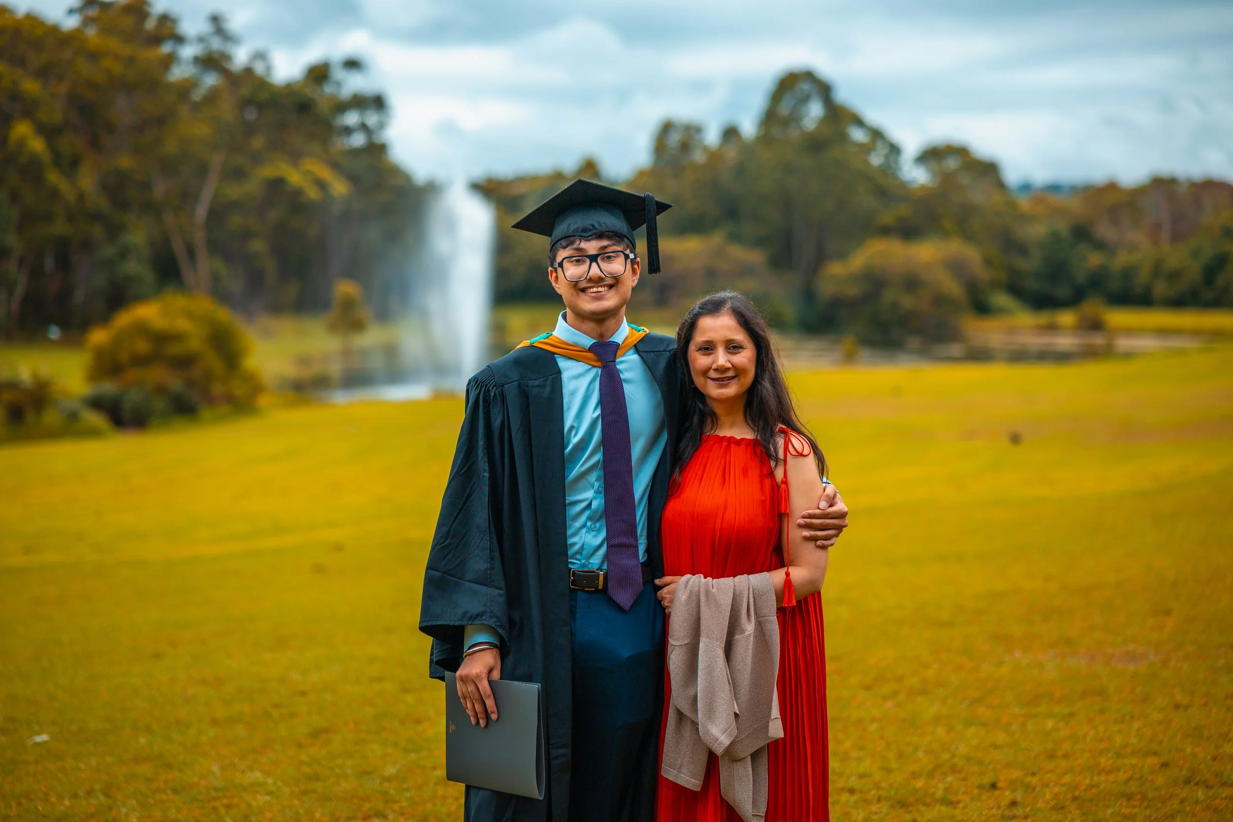 A young man in a graduation gown and cap standing next to a woman in a red dress in a park with a waterfall in the background.
