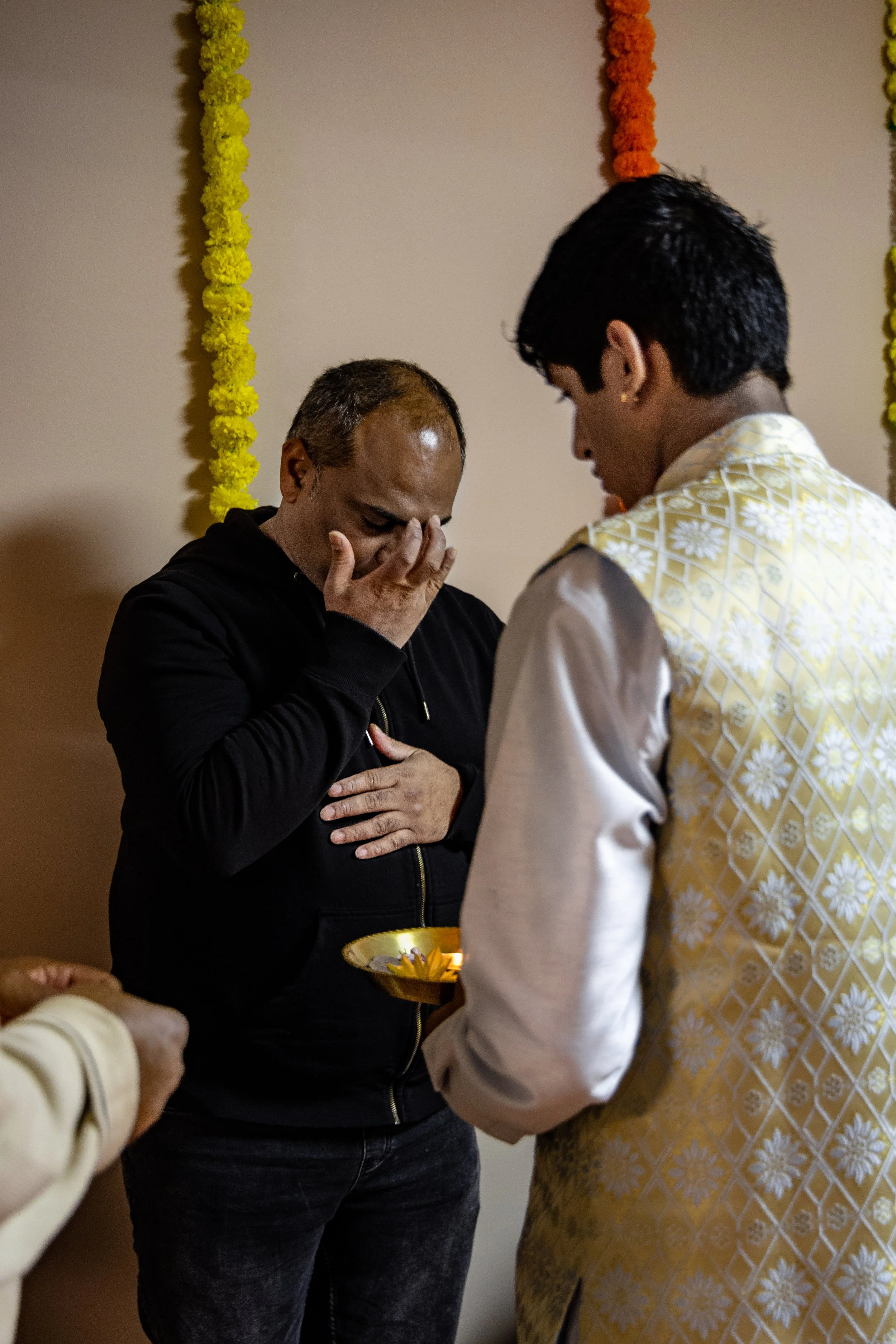 A man in black with a hand over his face appears to be praying during a religious ceremony as another man in traditional clothing offers a lit oil lamp in front of him, with colorful flower garlands hanging on the wall in the background.