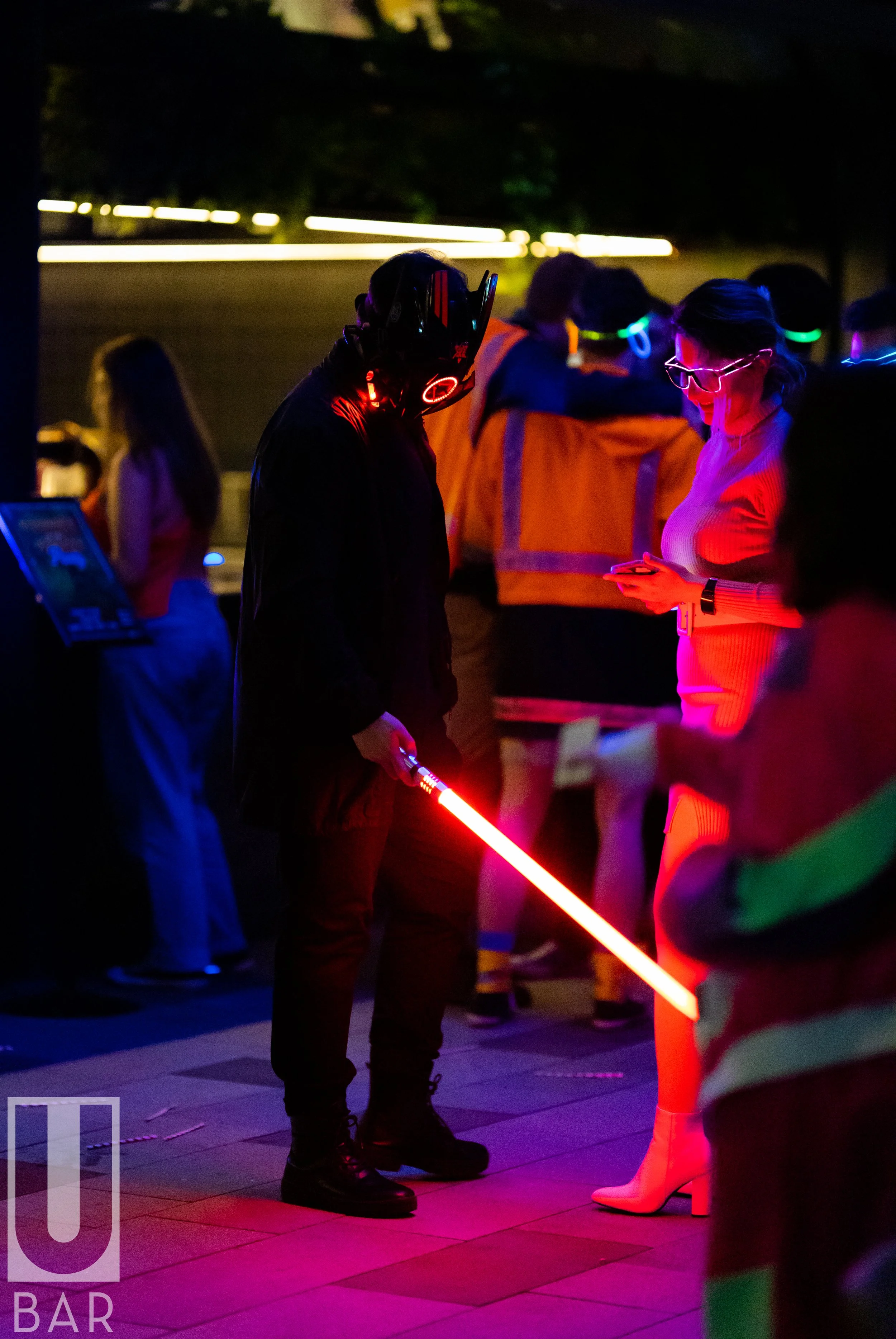 A person dressed as Darth Vader holding a red lightsaber in a nightclub or entertainment venue with several people around, some looking at their phones, illuminated by colorful lights.