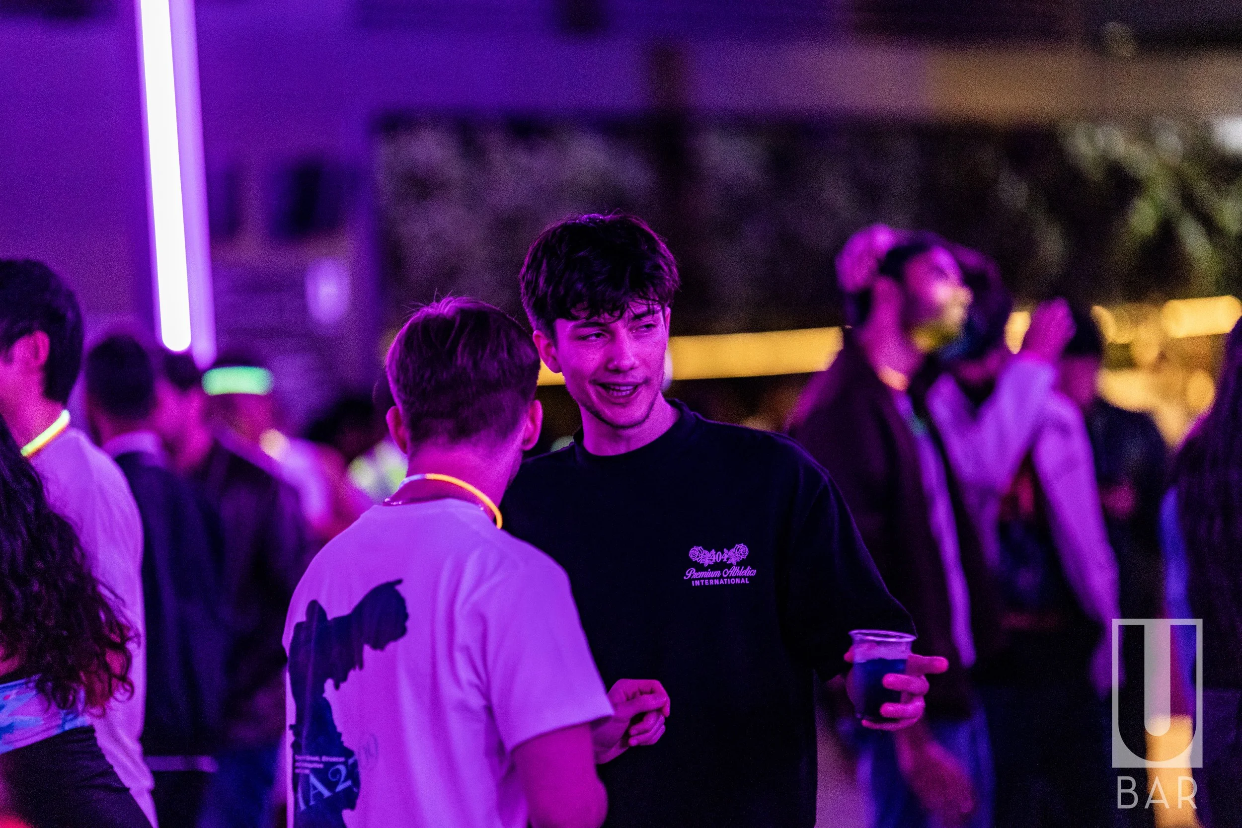 Two young men talking at a vibrant, neon-lit nighttime event with other attendees in the background, one holding a drink.