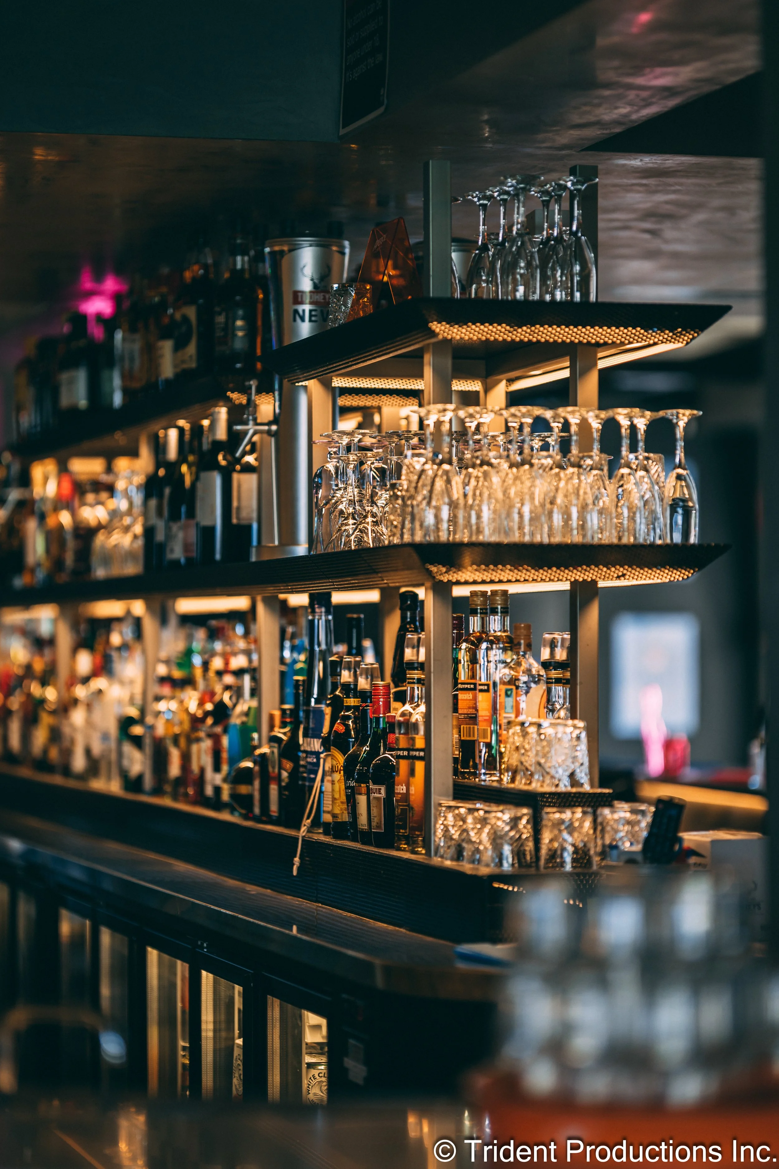 A bar with shelves stocked with various liquor bottles and upside-down glasses, illuminated by warm lighting.