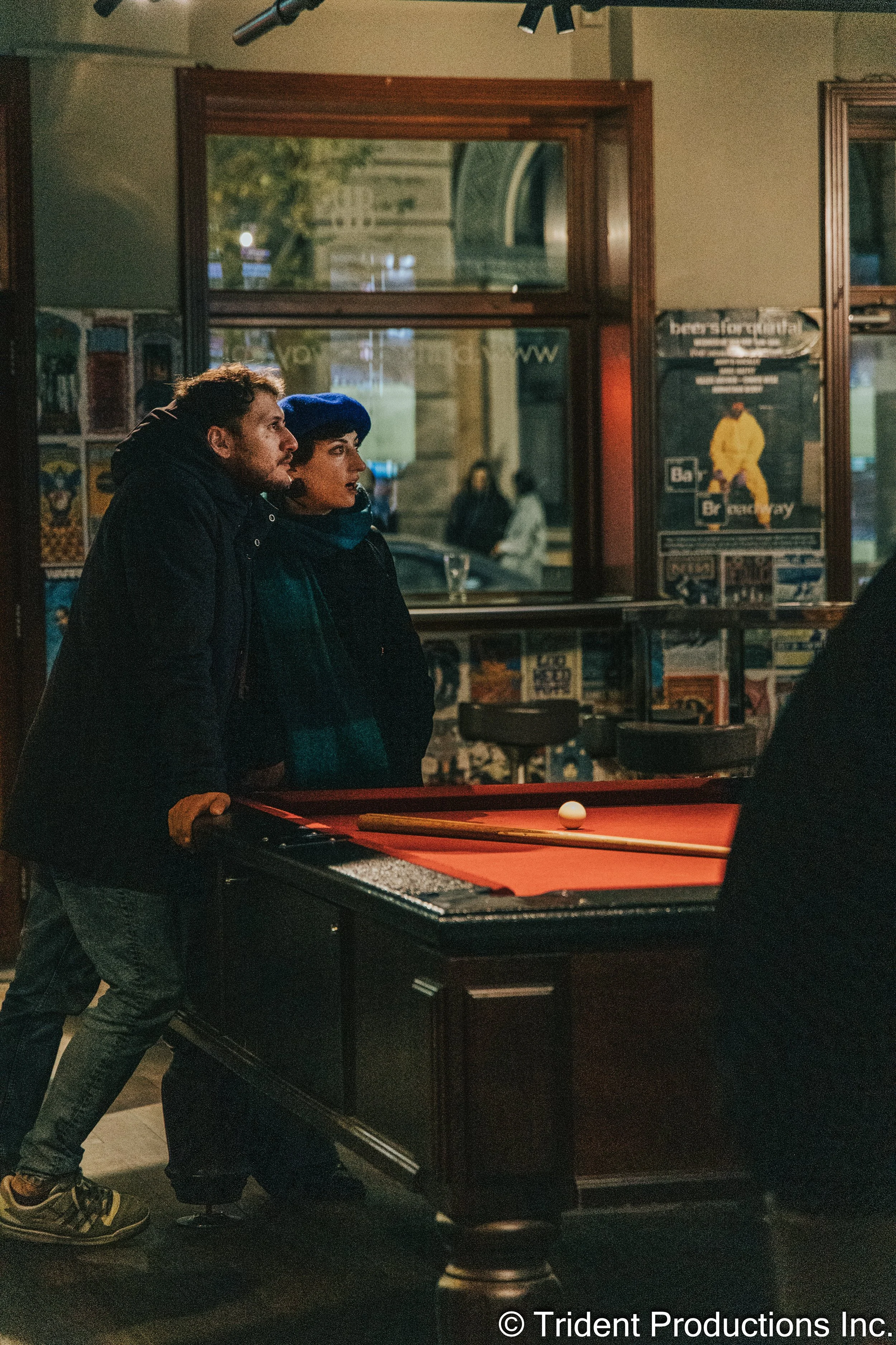 A man and woman standing together at a pool table in a dimly lit bar, watching the game. The man is leaning on the table, and the woman is wearing a blue beret. There are posters on the wall behind them and a window showing a street scene outside.