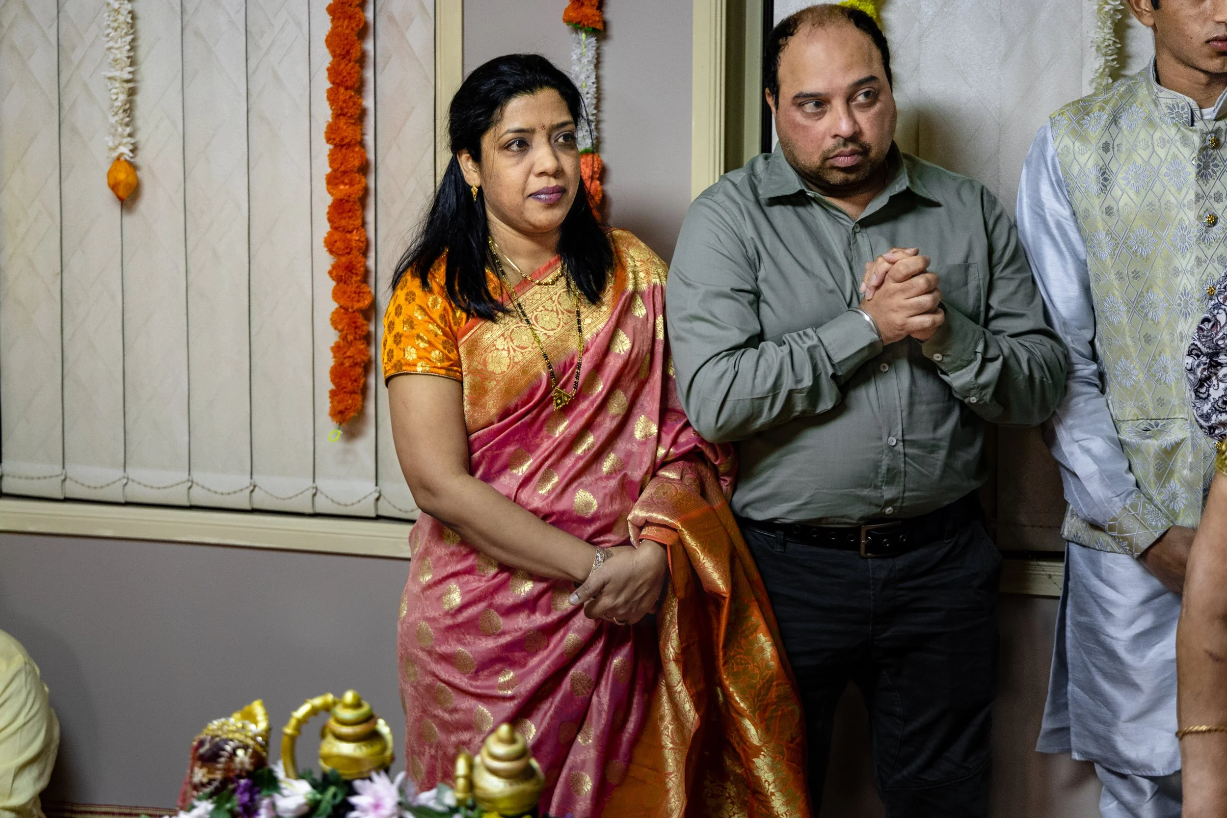 Indian woman in a traditional pink and gold sari and a man in a gray shirt praying with hands clasped during a cultural or religious event, decorated with garlands and flowers.