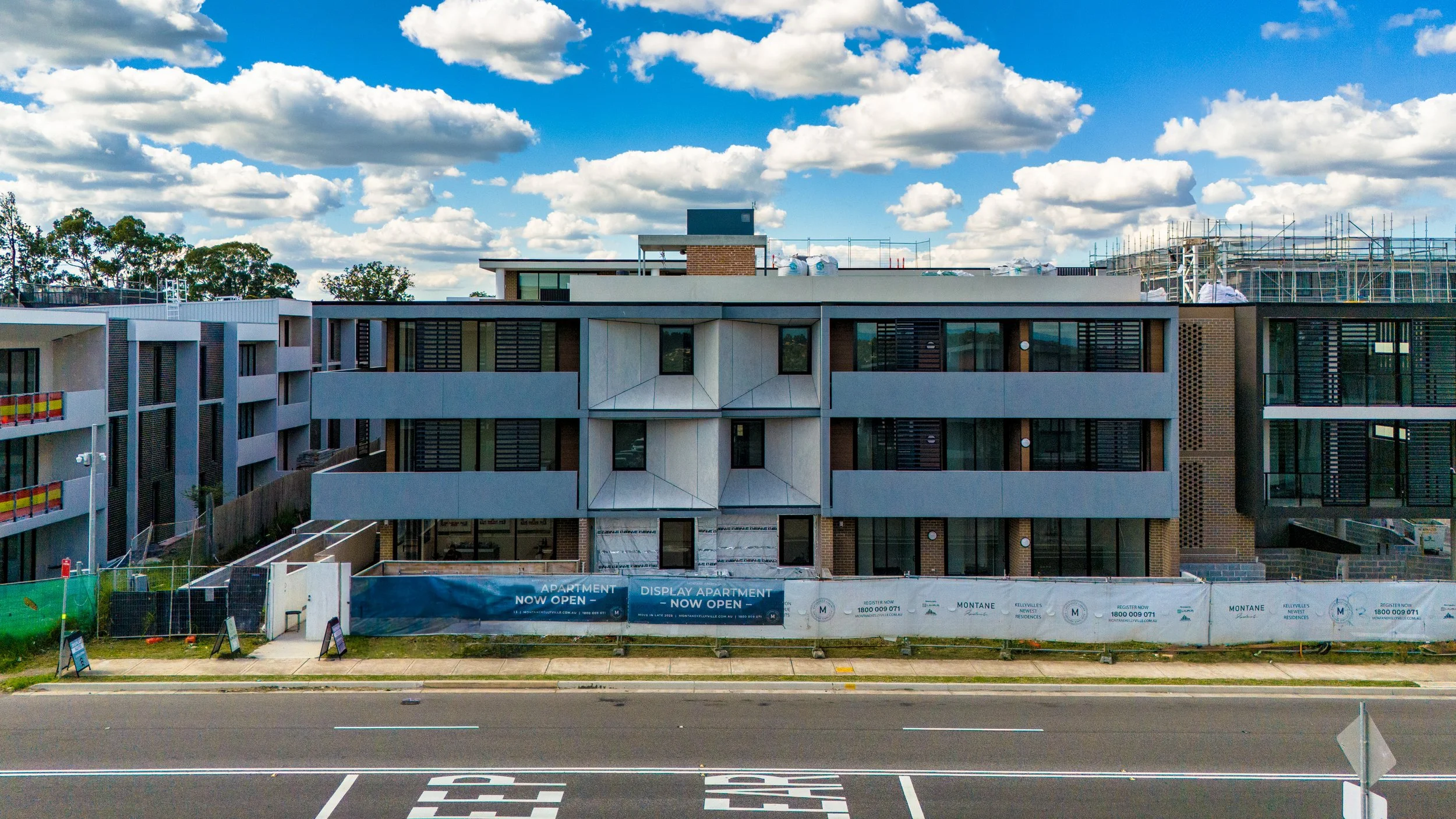 Under construction modern apartment building with multiple floors, large windows, and balconies, with a blue sky and white clouds in the background, and a road with marked lanes in the foreground.
