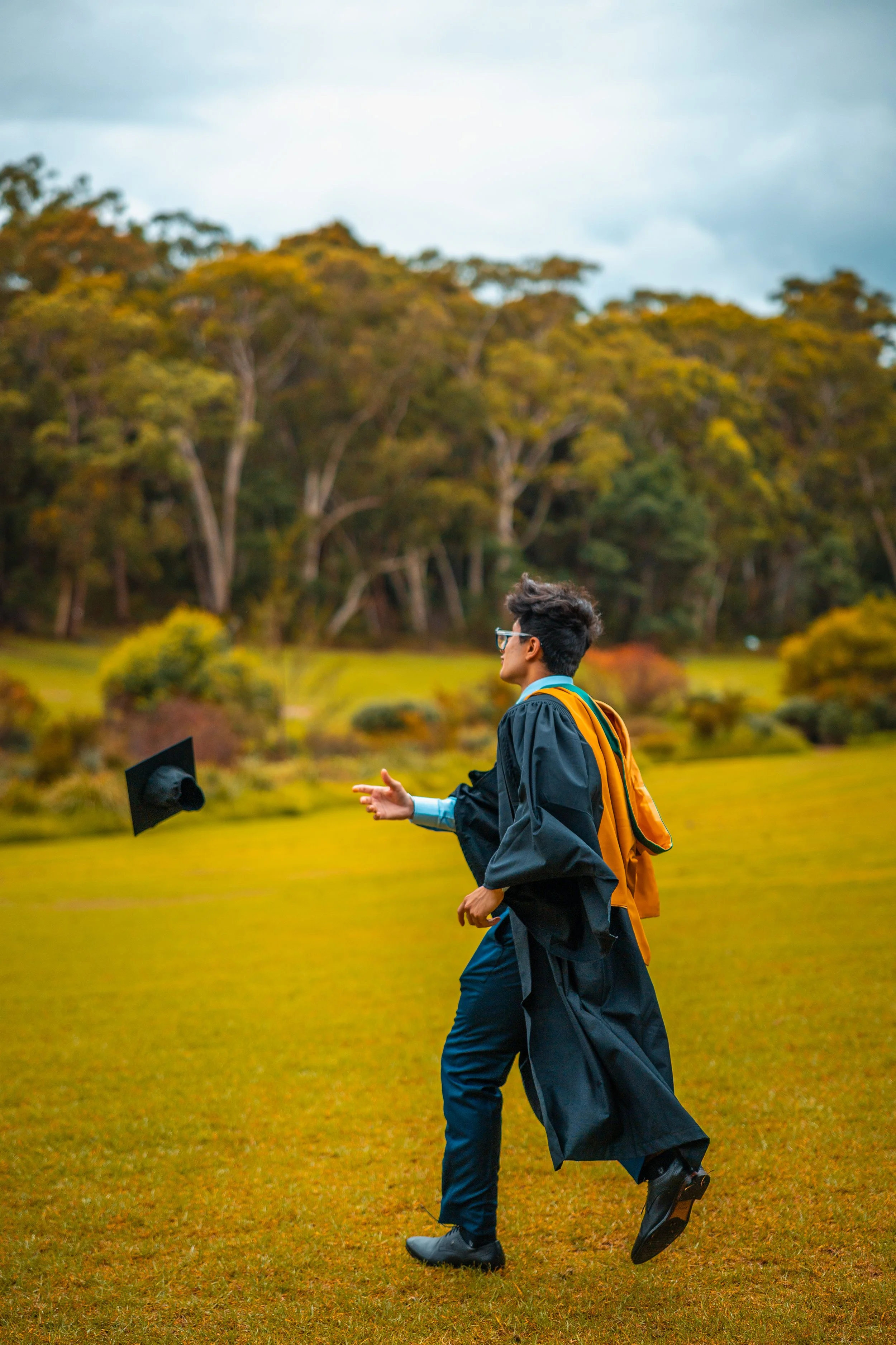 A young man in a graduation gown and cap throwing his cap in the air in a grassy outdoor setting with trees in the background.