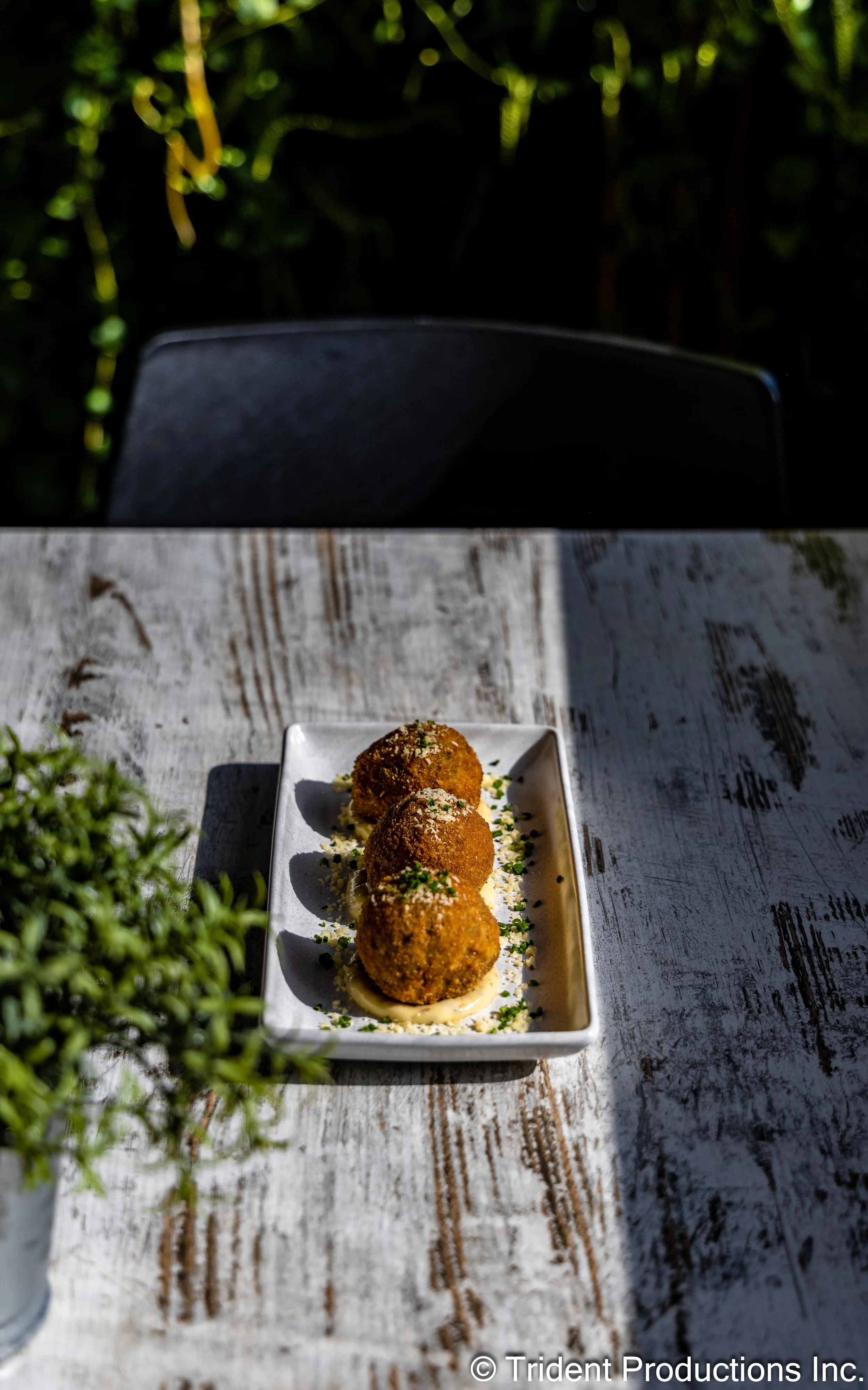 Three fried arancini balls served on a rectangular white plate with garnished herbs on a rustic white wooden table outside.
