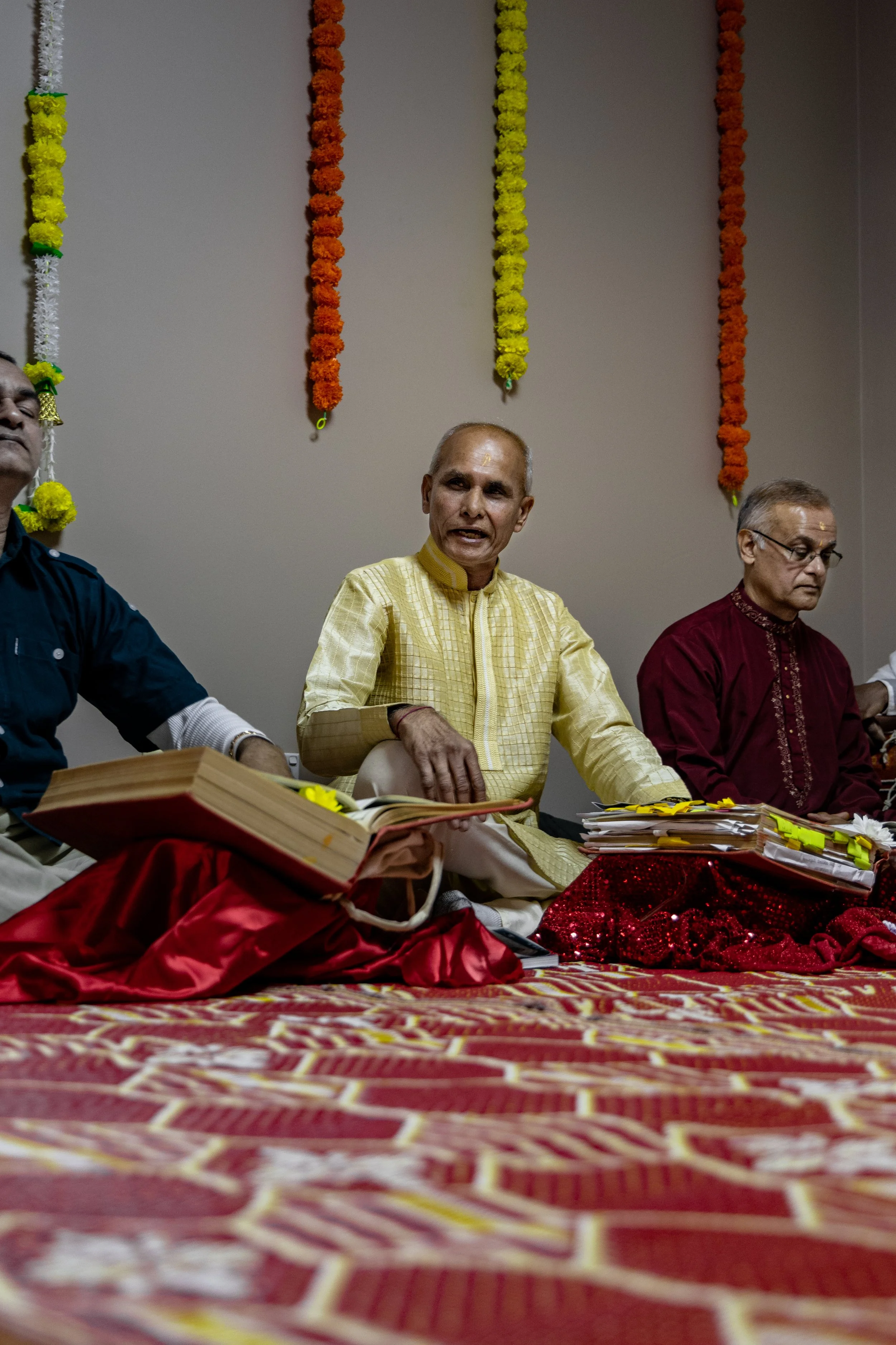 Three men sitting cross-legged on a decorated floor, participating in a traditional ceremony, with colorful flower garlands hanging on the wall behind them.