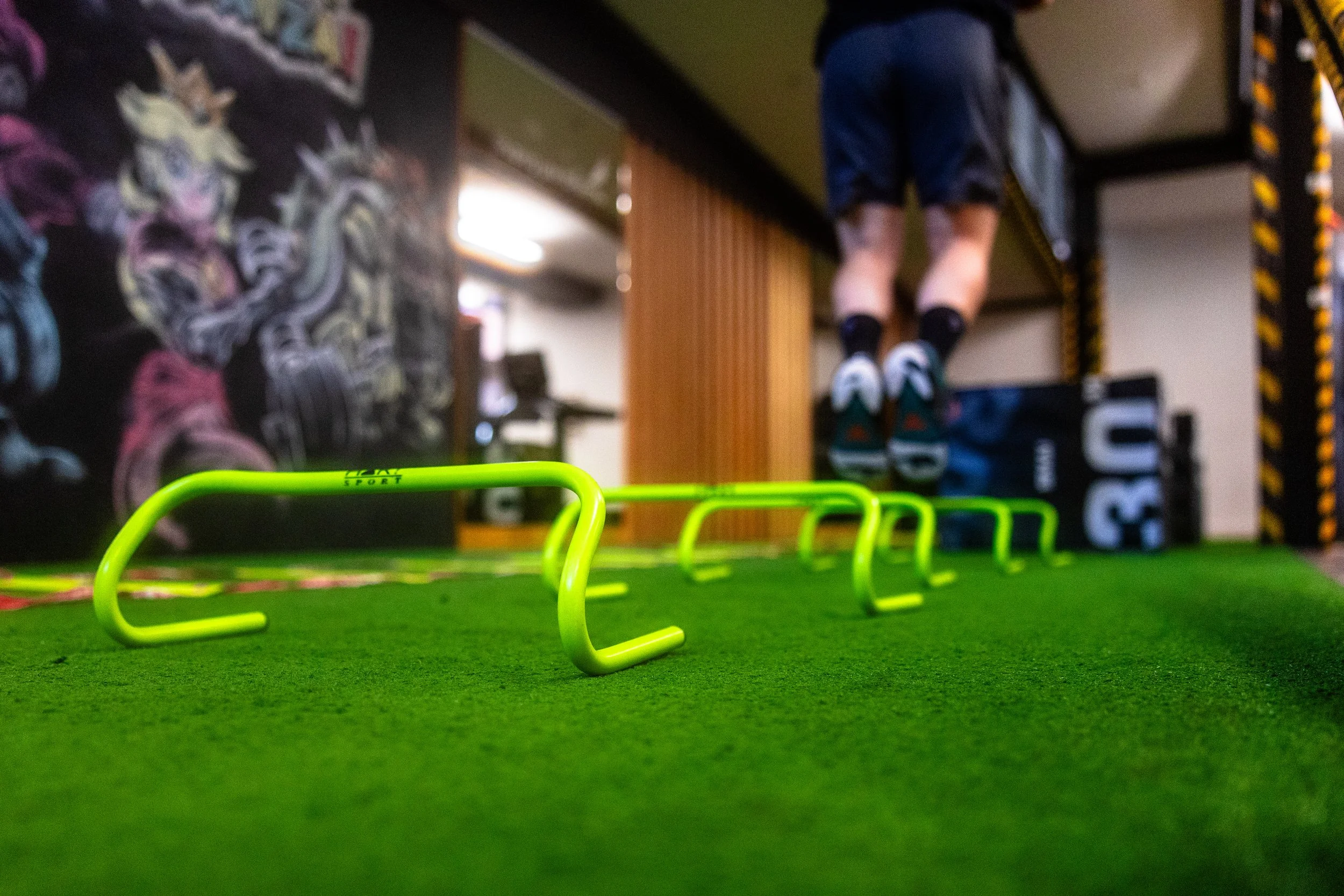 Close-up of green workout hurdles on artificial green turf, with a person in athletic shorts and sneakers jumping on a plyometric box in the background of a gym.