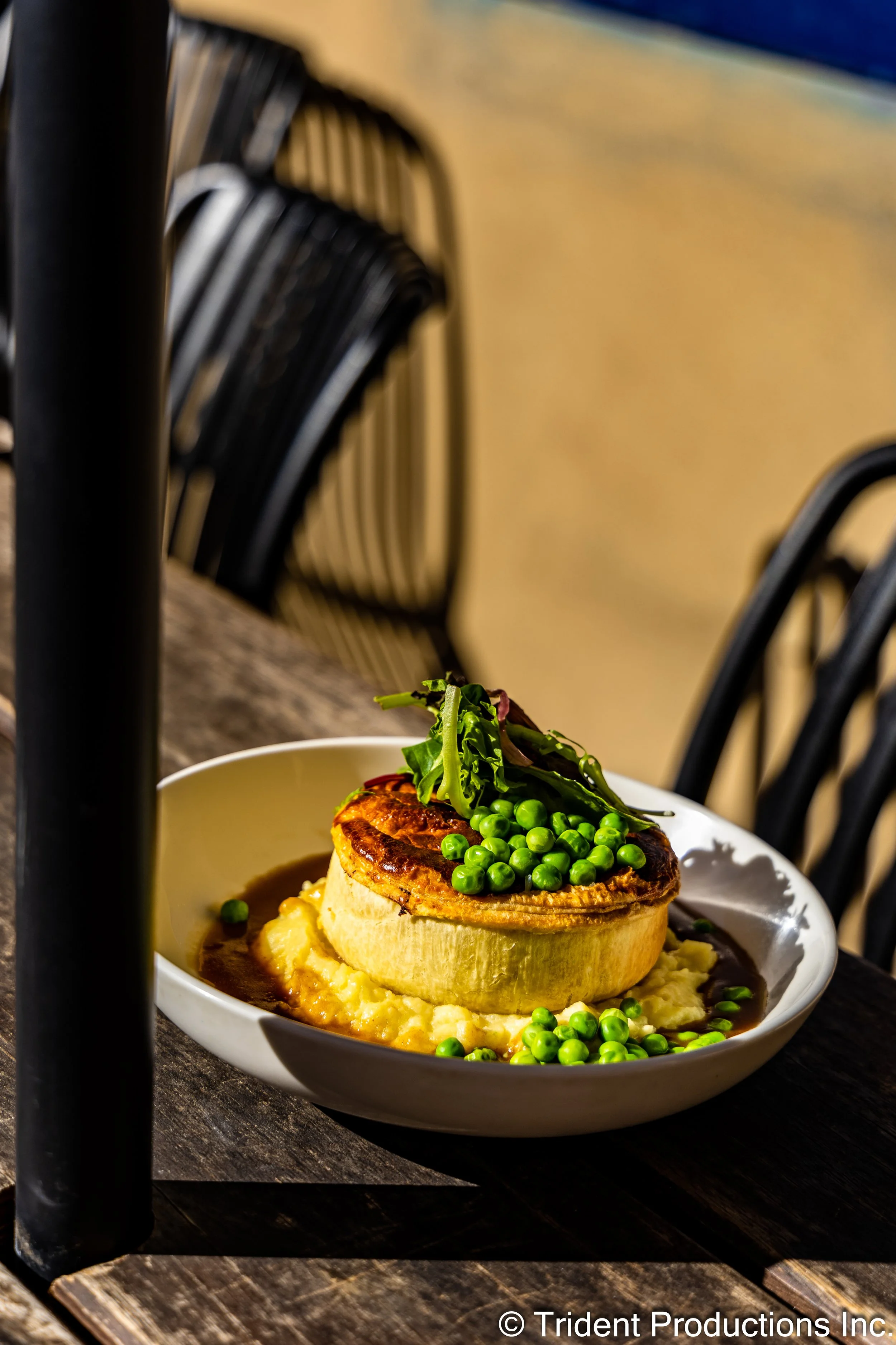 A plated dish featuring a large round piece of meatloaf with a brown crust, topped with fresh greens and green peas, served over mashed potatoes with gravy, on a white oval plate.