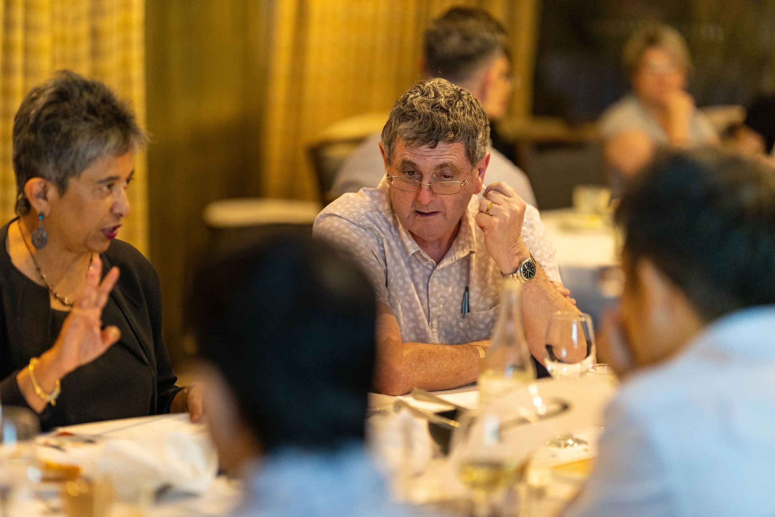 People sitting around a dining table engaged in conversation, with one woman gesturing and a man wearing glasses listening.