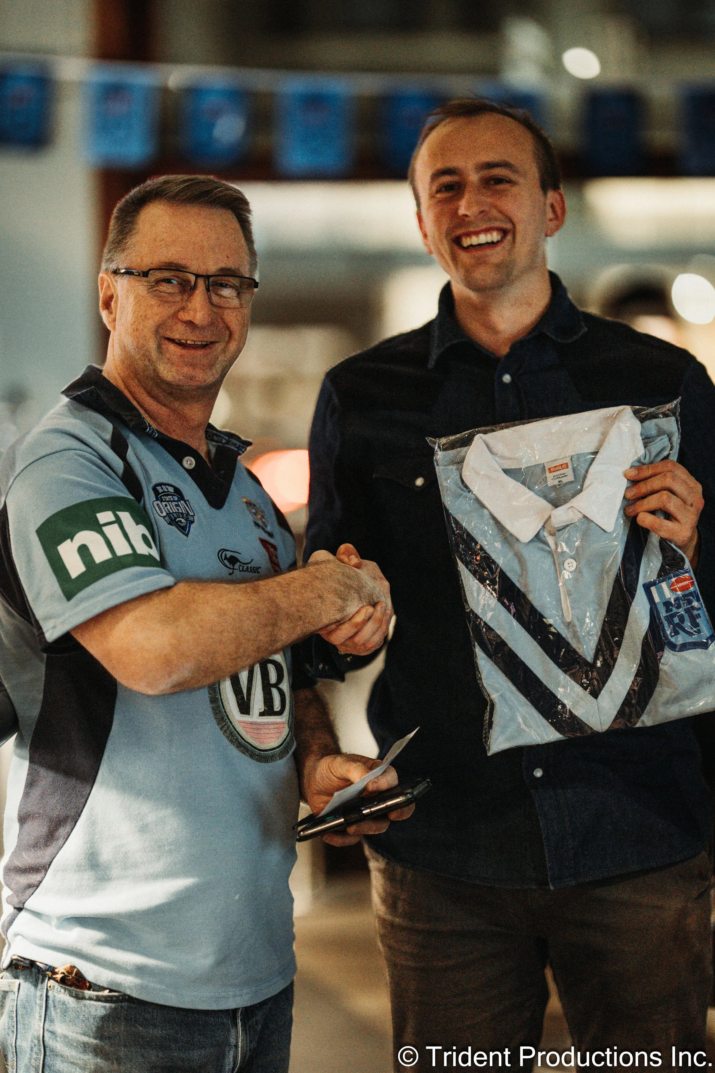 Two men shaking hands, one wearing a light blue sports jersey and the other holding a packaged collared shirt. They are smiling at an indoor event.