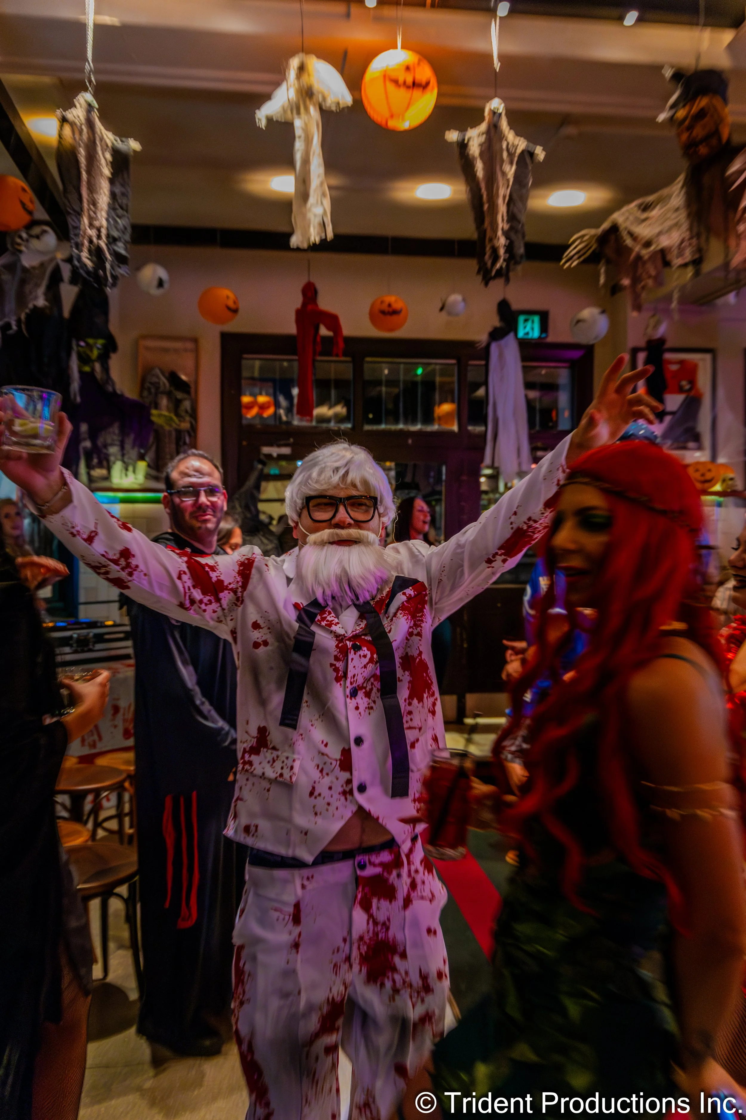 People in Halloween costumes celebrating at a party with Halloween decorations, including hanging ghosts, pumpkins, and a red cloth hanging from the ceiling.