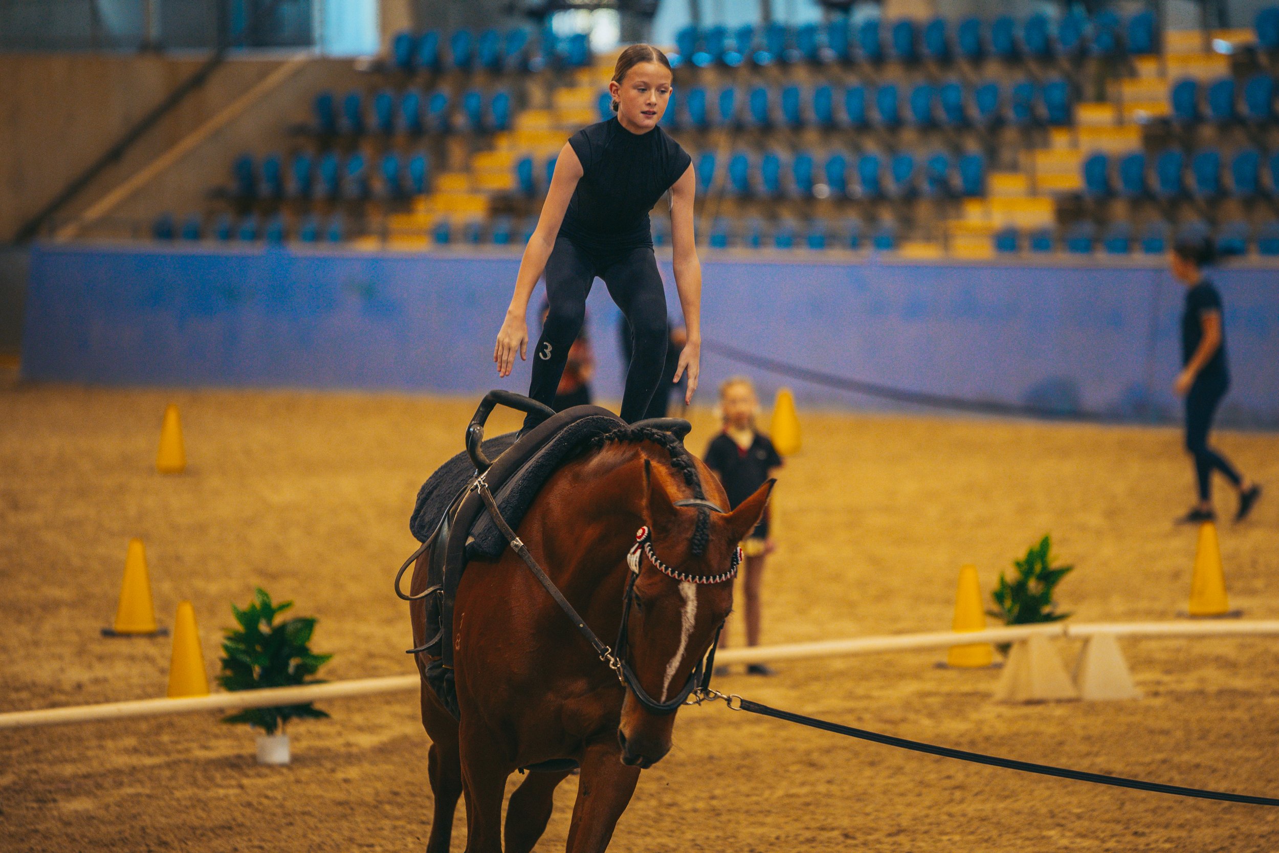 A young girl practicing equestrian skills on a brown horse inside an indoor riding arena, with other people and yellow cones in the background.