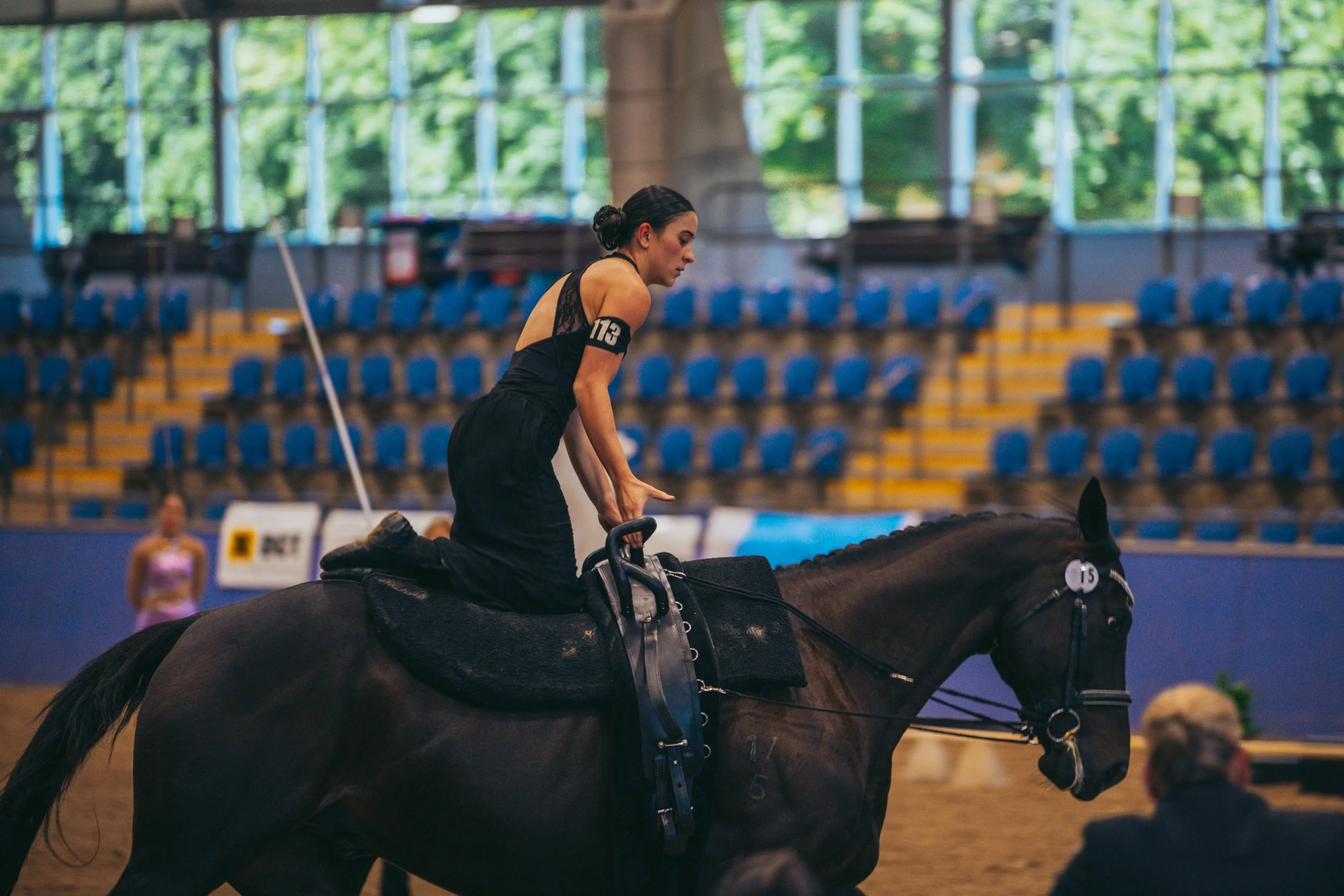 A woman riding a horse in an indoor arena. She is wearing black attire and a numbered armband, with a woman and a man in the background.