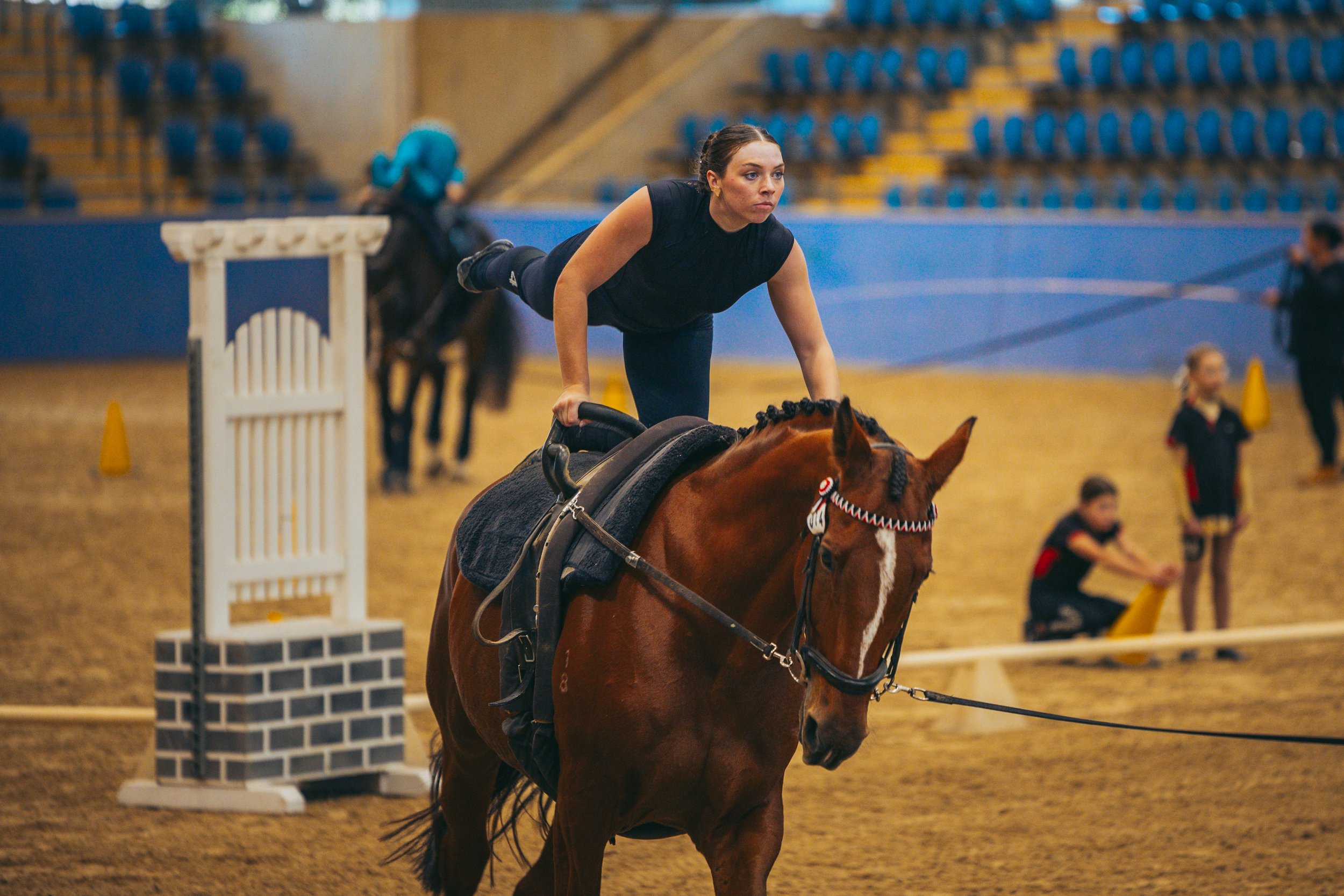 A woman riding a horse in an indoor arena, practicing show jumping. She is wearing athletic clothing and balancing on the horse as it approaches a small jump obstacle.