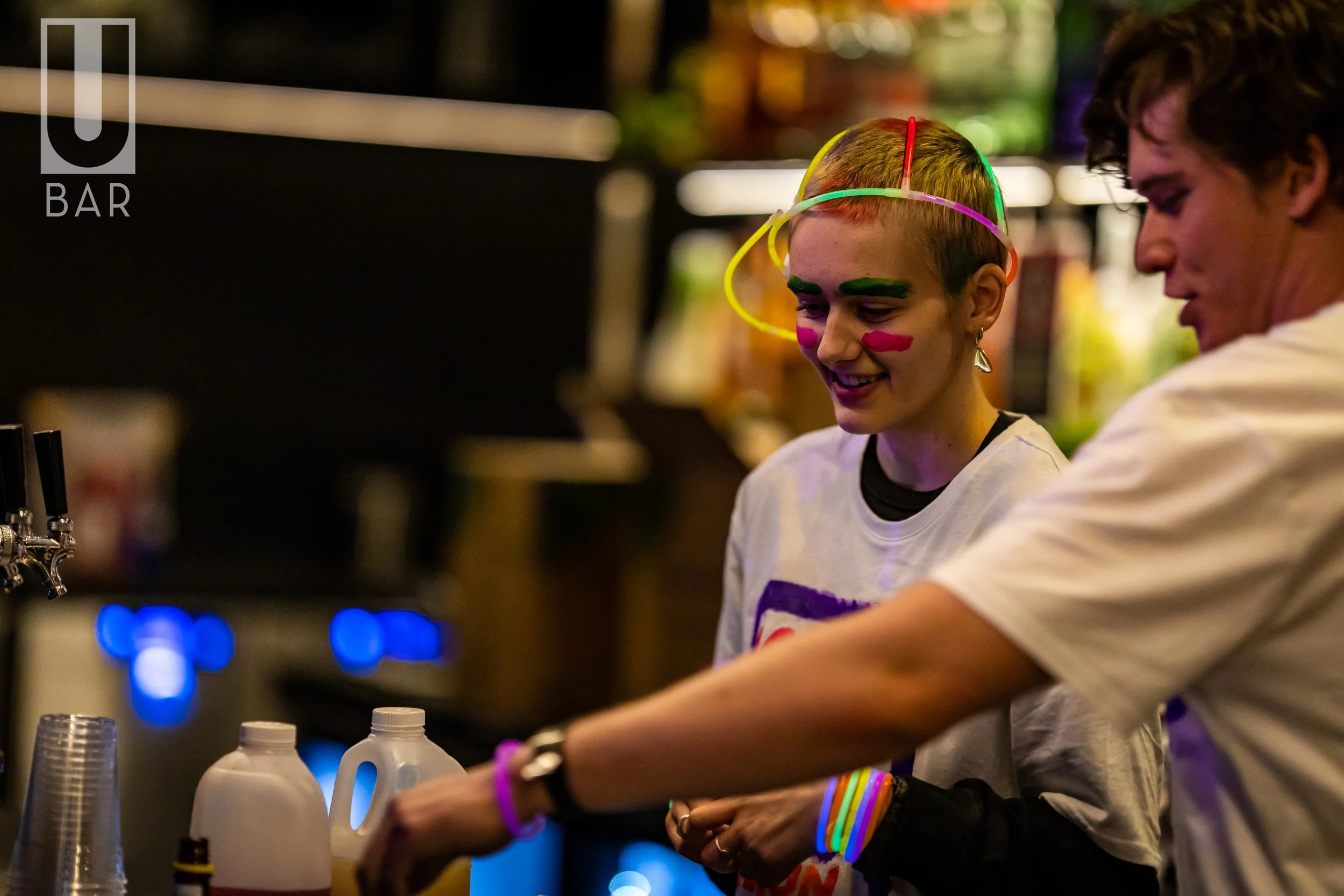 Two young people at a bar, one with neon colored glow sticks and face paint, both smiling, preparing drinks.